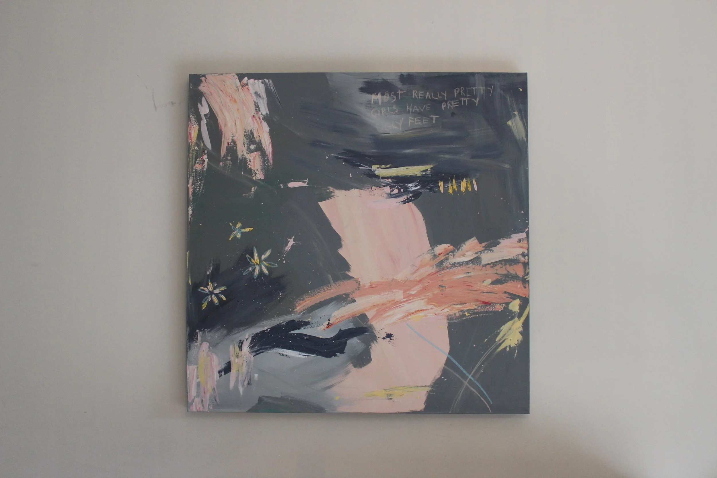 Abstract expressionist painting on a canvas with dark gray, pink, and yellow brushstrokes, some floral elements, and partial white text on a plain white wall.