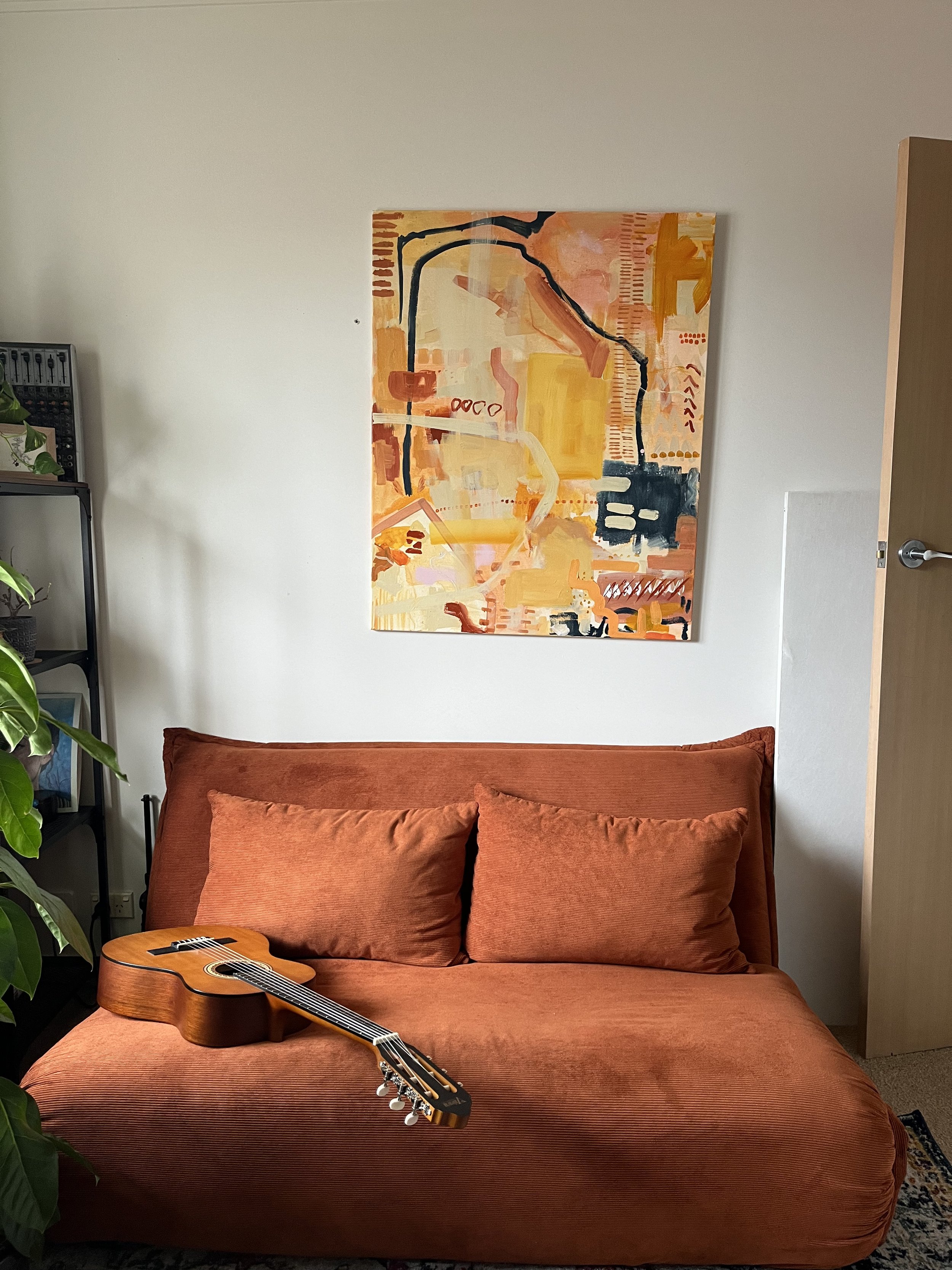 A room with a burnt orange couch, a guitar resting on it, a large abstract painting with warm colors on the wall, and some green plants on the left side.