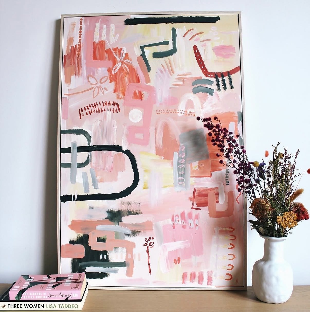An abstract colorful painting with pink, red, black, gray, yellow, and white strokes, next to a white vase with a bouquet of dried flowers and a pink book titled 'Three Women' by Lisa Taddeo.