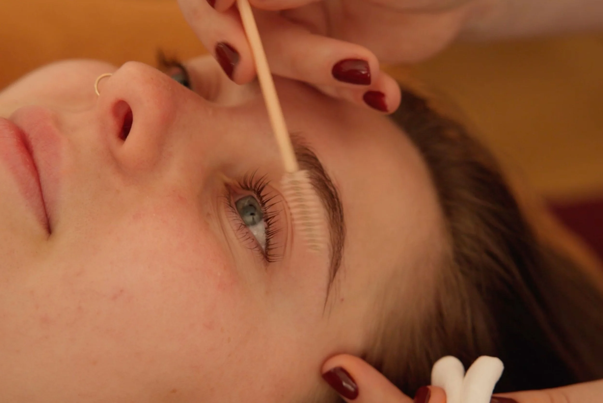 A person with light skin, light blue eyes, and dark red nail polish on their fingers is receiving dental care, including the use of a dental mirror and dental tools. The person's face is relaxed, and they are lying down with their mouth open during an examination.