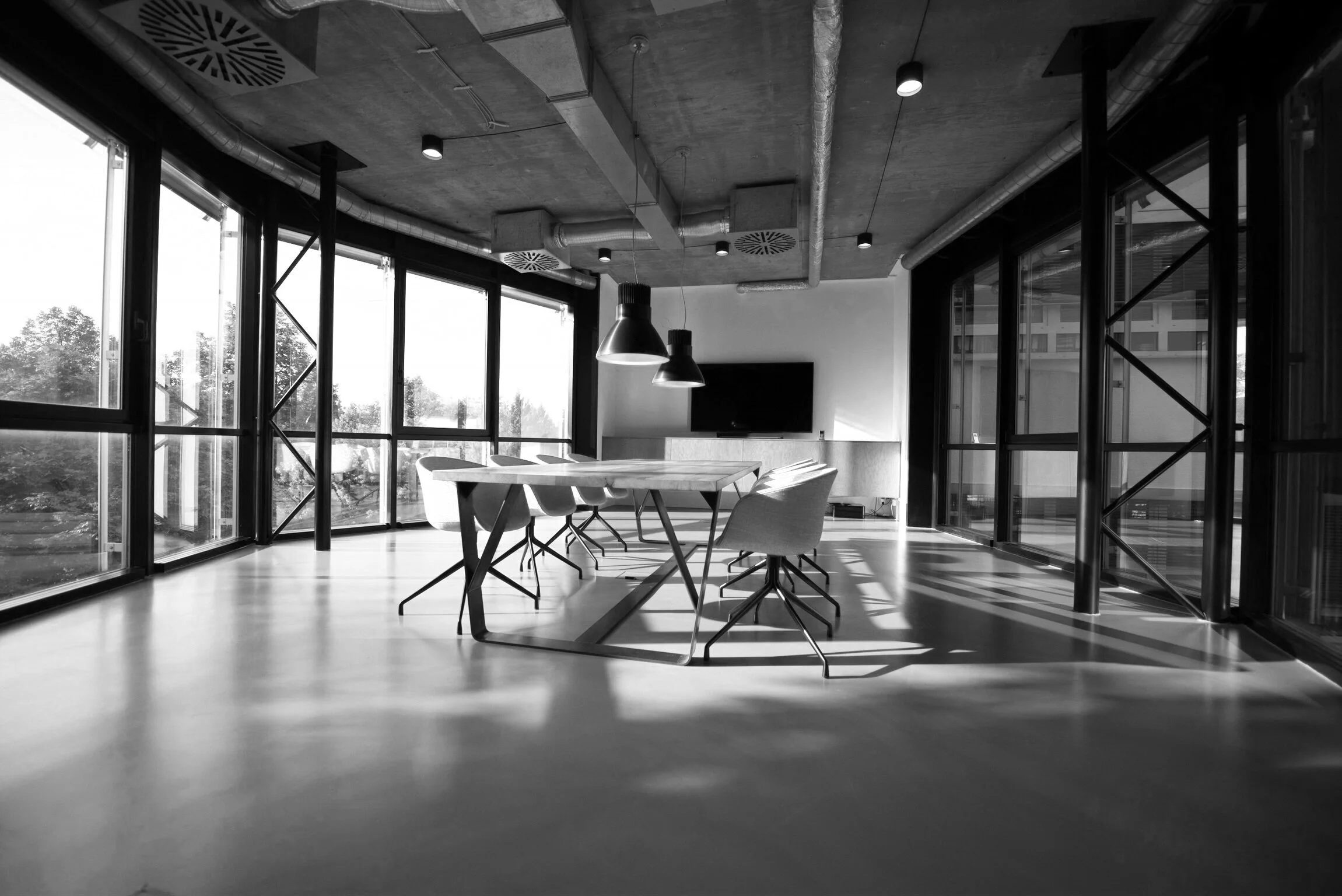 Modern conference room with large windows, a rectangular table, six chairs, pendant lights, and a wall-mounted TV, with sunlight casting shadows inside.