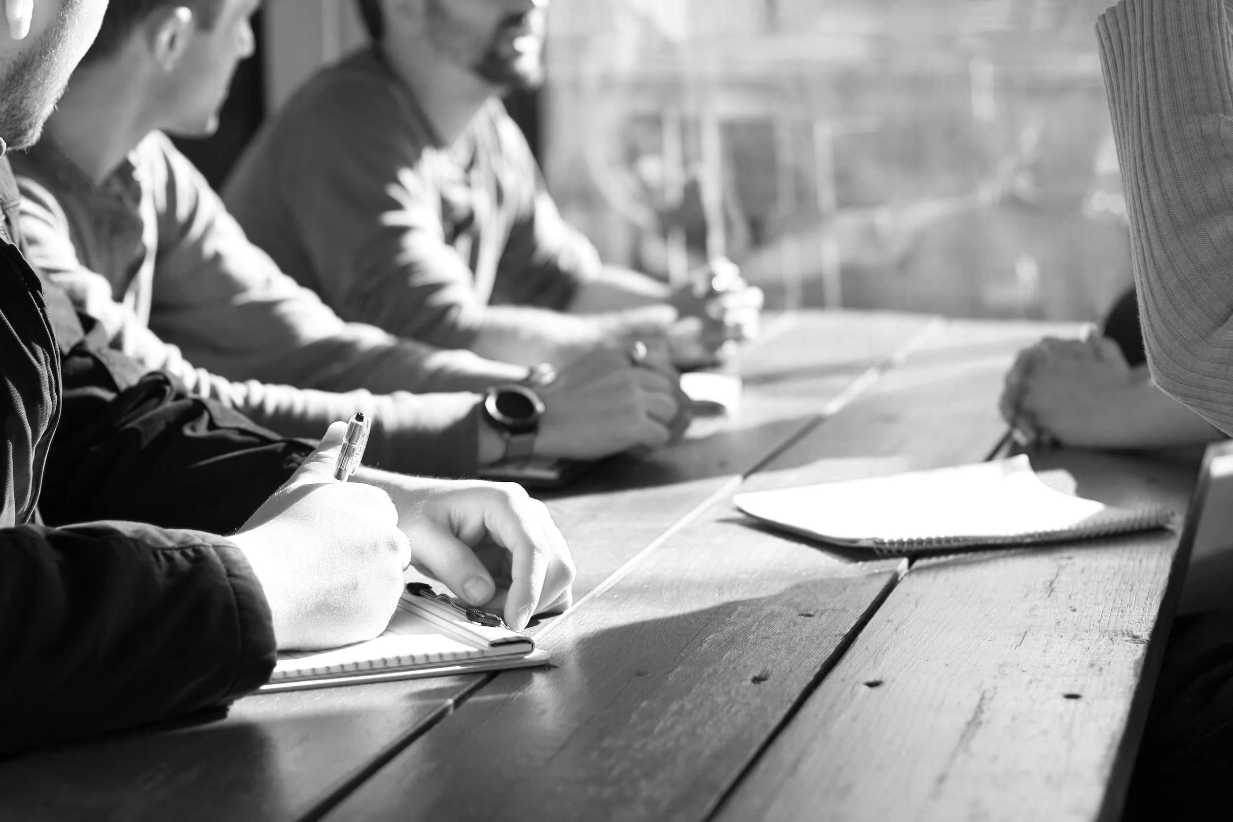 People sitting at a wooden table taking notes and writing, in a meeting or discussion, sunlight coming through a window.