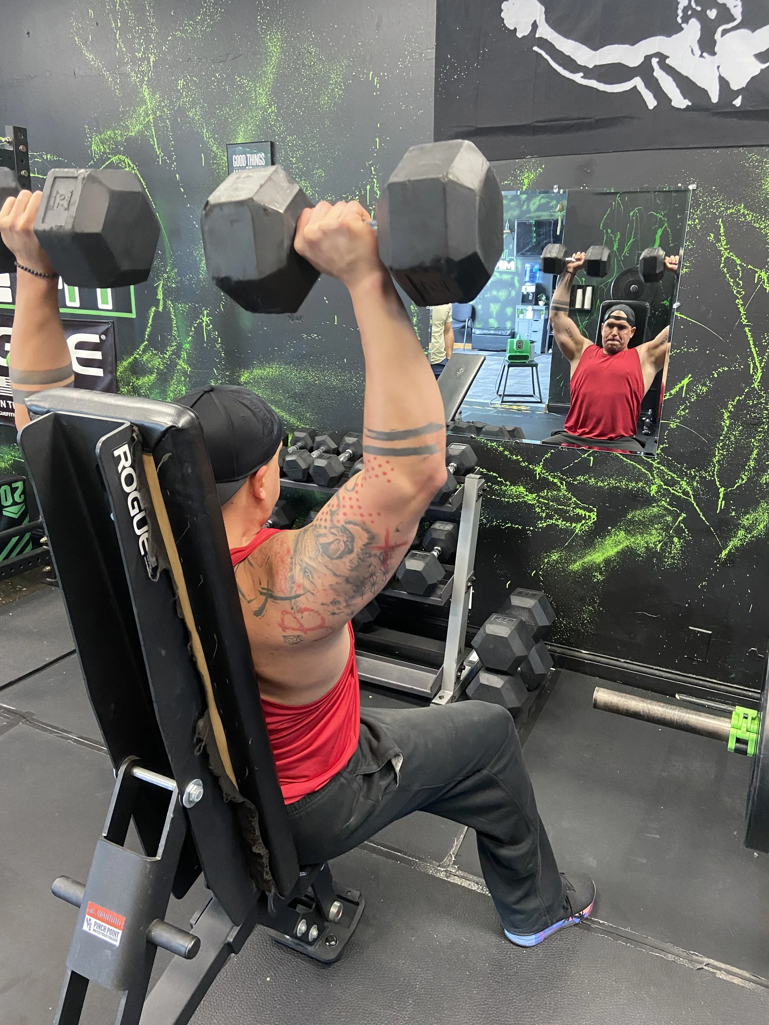 A man with tattoos wearing a red tank top and a black cap is seated on a vertical workout bench, lifting two dumbbells with both arms. His reflection is visible in a mirror on the wall behind him. The gym has black walls with green splatter art and workout equipment.