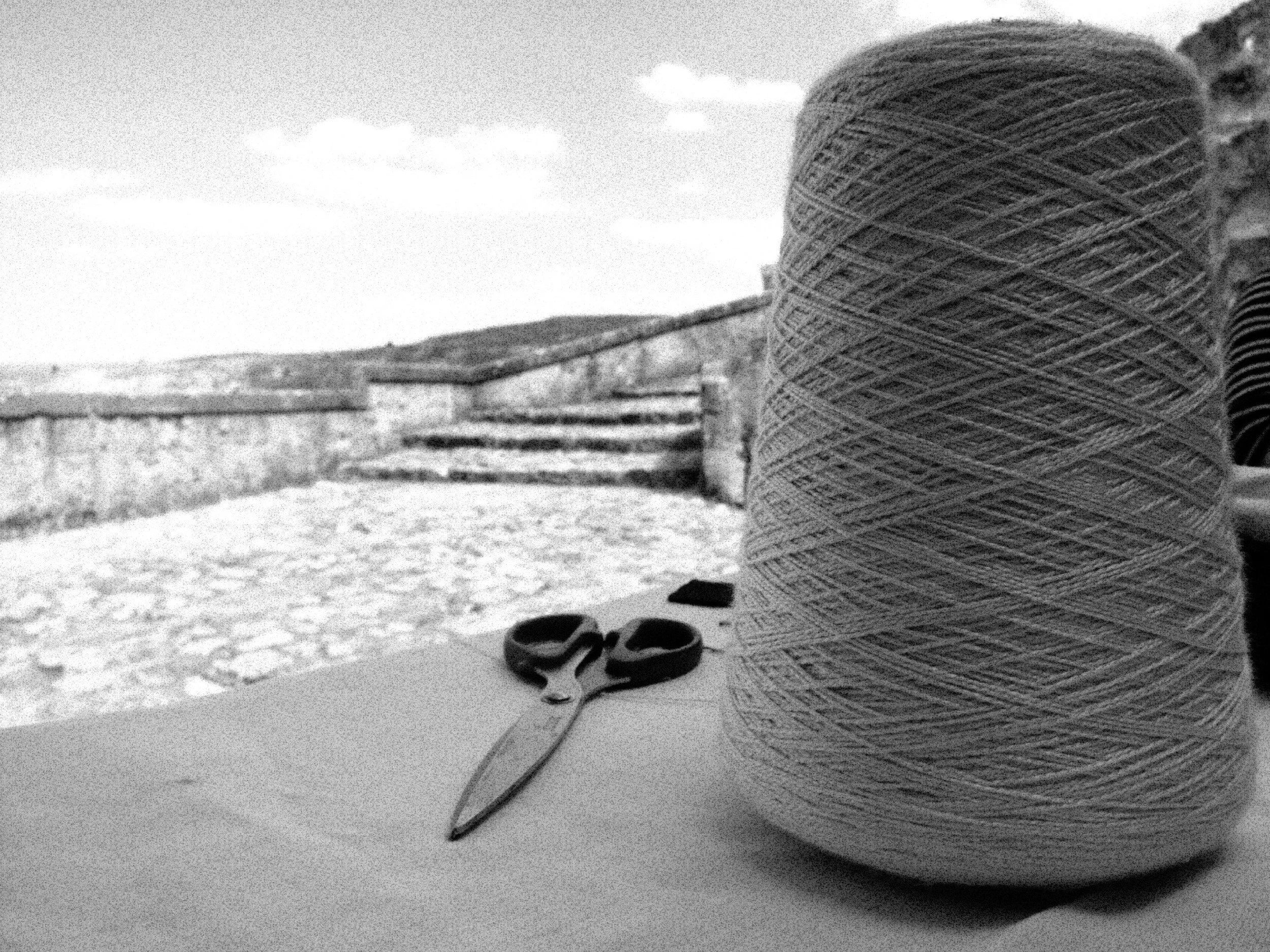 A ball of yarn with knitting needles and scissors on a flat surface with a house and hills in the background.