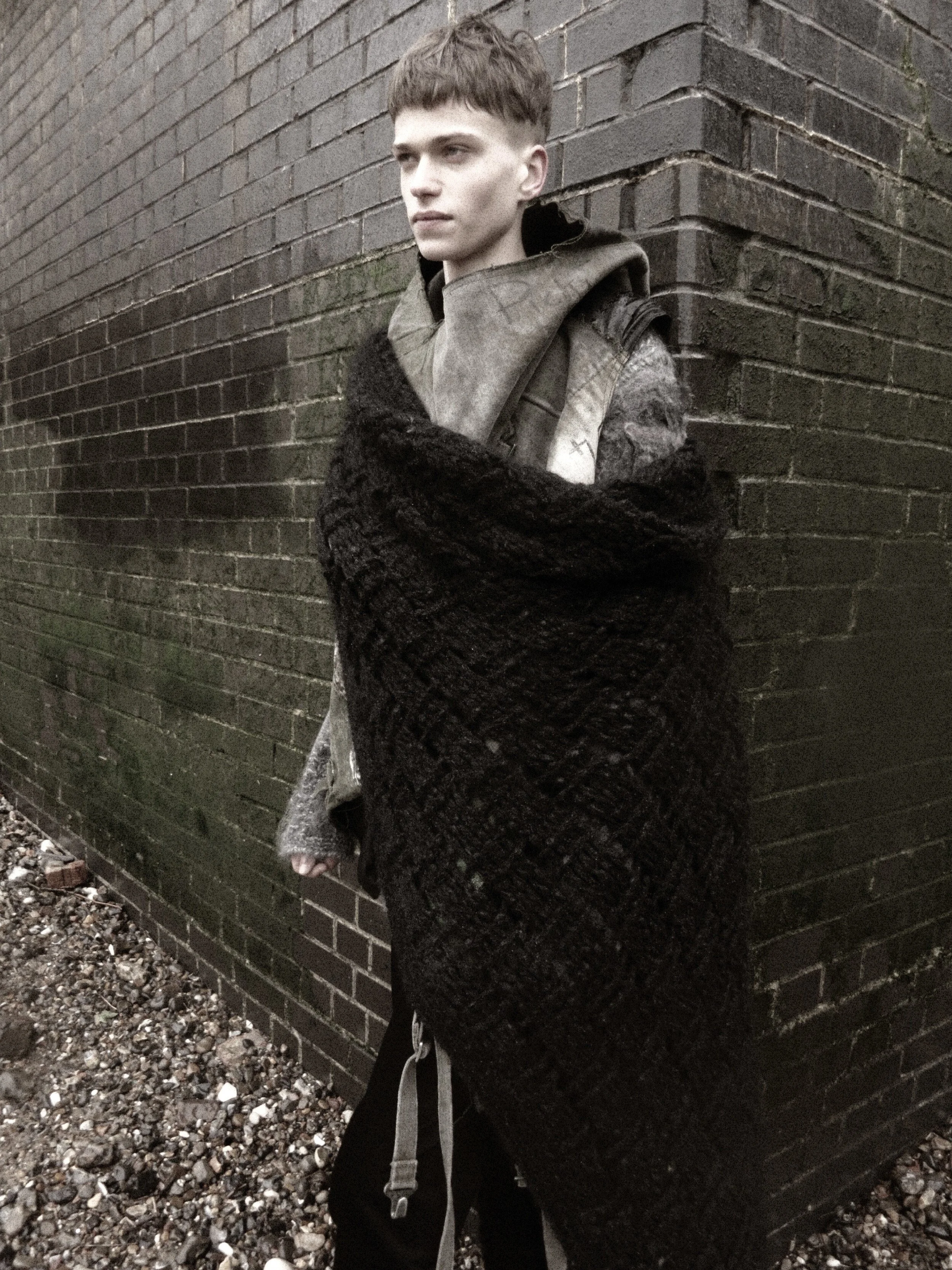 A young person leaning against a dark brick wall, wearing a black and grey sweater, with short hair, looking thoughtfully into the distance.