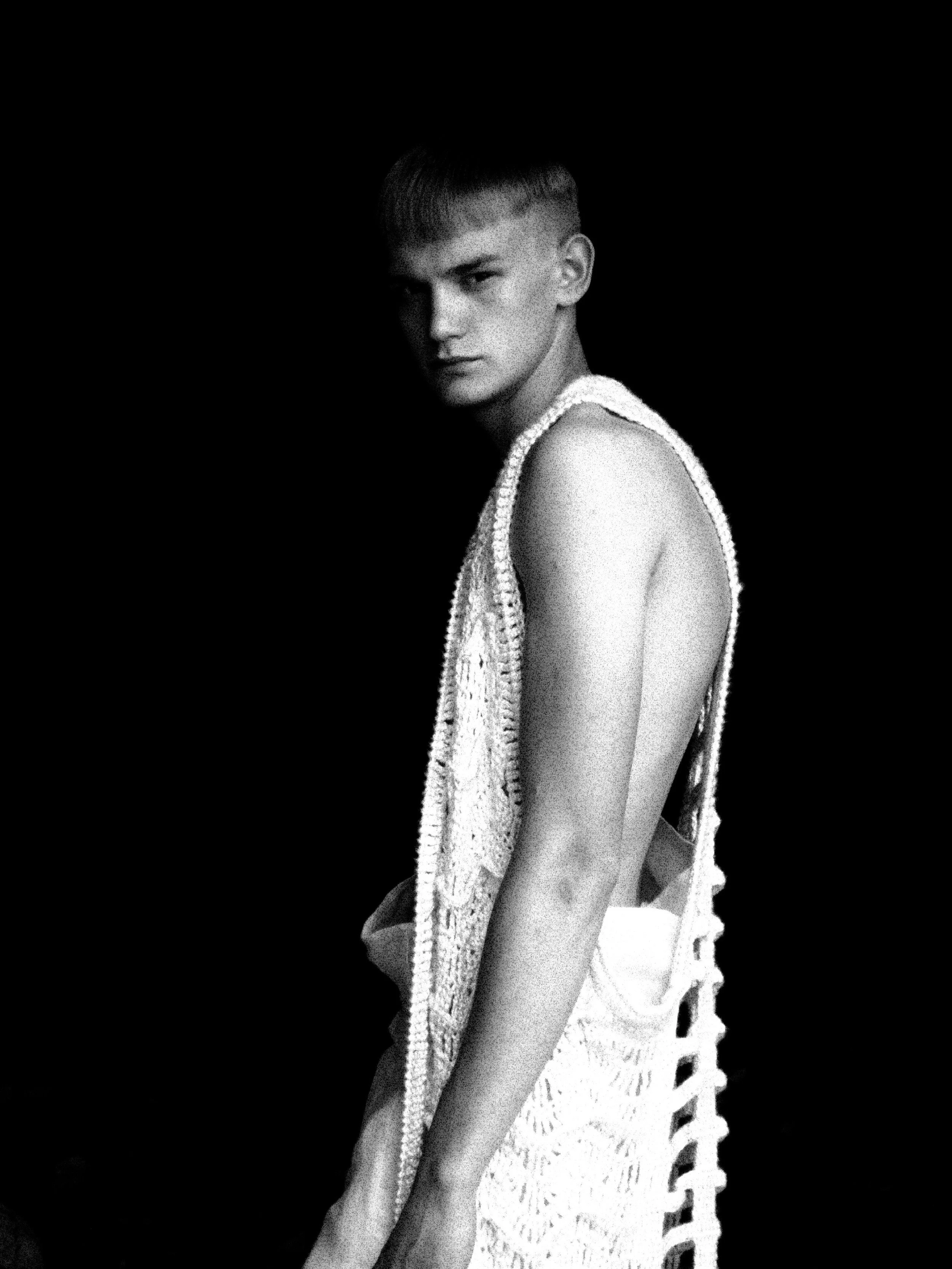 Black and white photo of a young woman with short hair, wearing a sleeveless knitted top, looking seriously at the camera against a dark background.