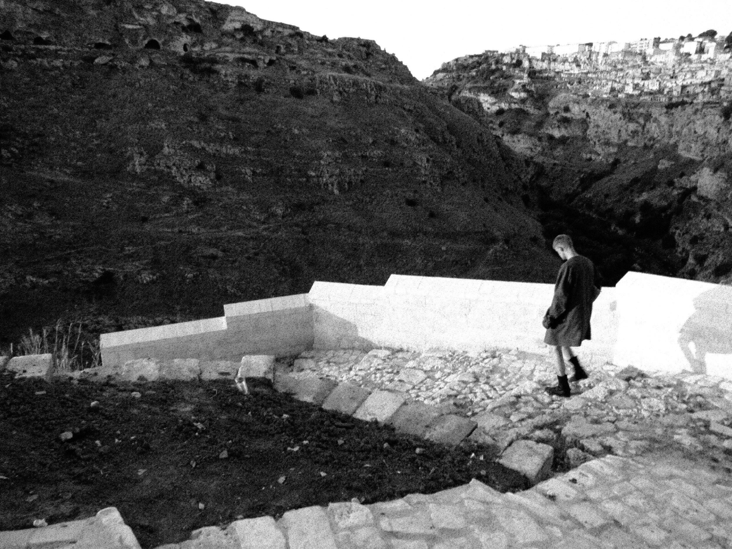 A black-and-white photo showing two people standing outdoors near a staircase made of stone steps. The background features a rugged hillside with rocky terrain and sparse vegetation, and a row of buildings atop the hillside.
