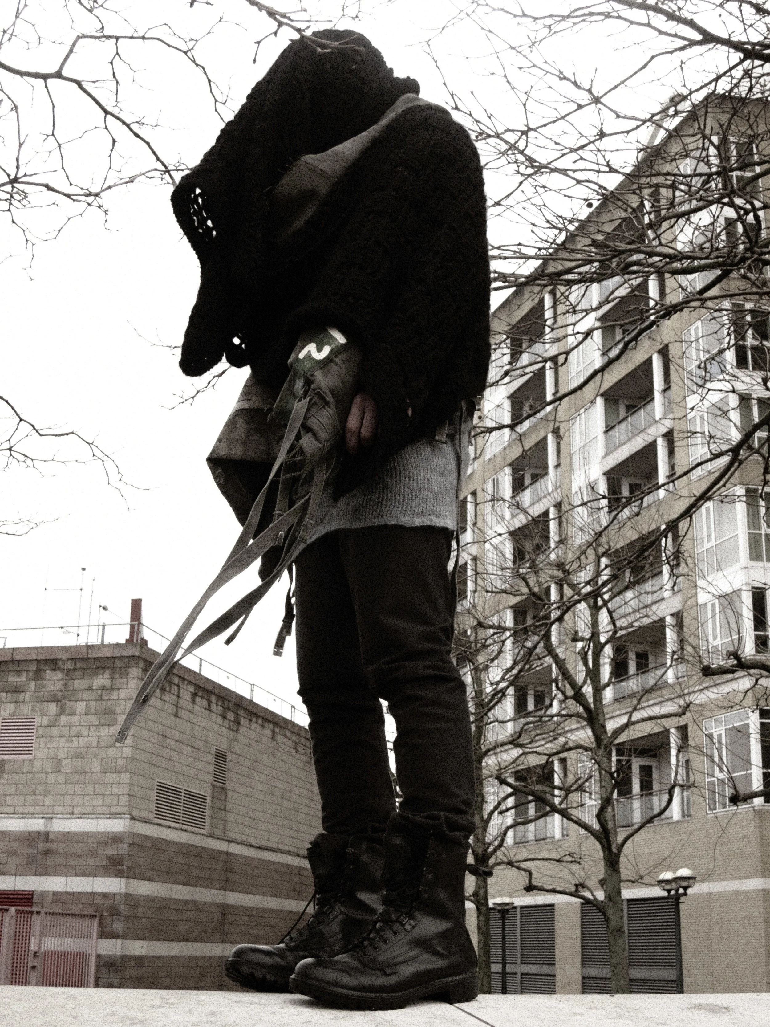 A person standing outdoors in an urban area, wearing large hiking boots, gray pants, a dark jacket, and a gray sweater, holding a camera and looking upwards. There is a building with multiple windows and leafless trees in the background during daytim
