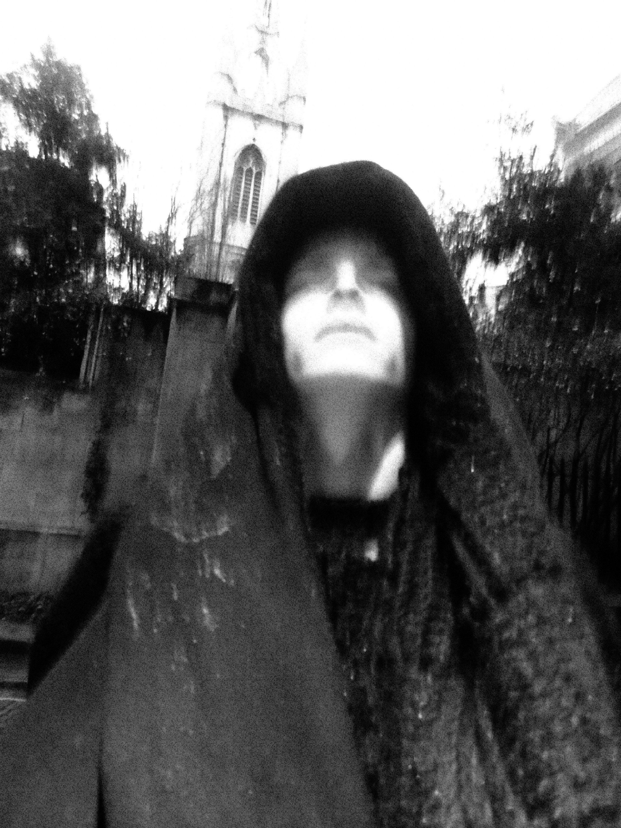 A black and white photo of a person with short hair and a hooded jacket, standing outside in front of a church and trees.