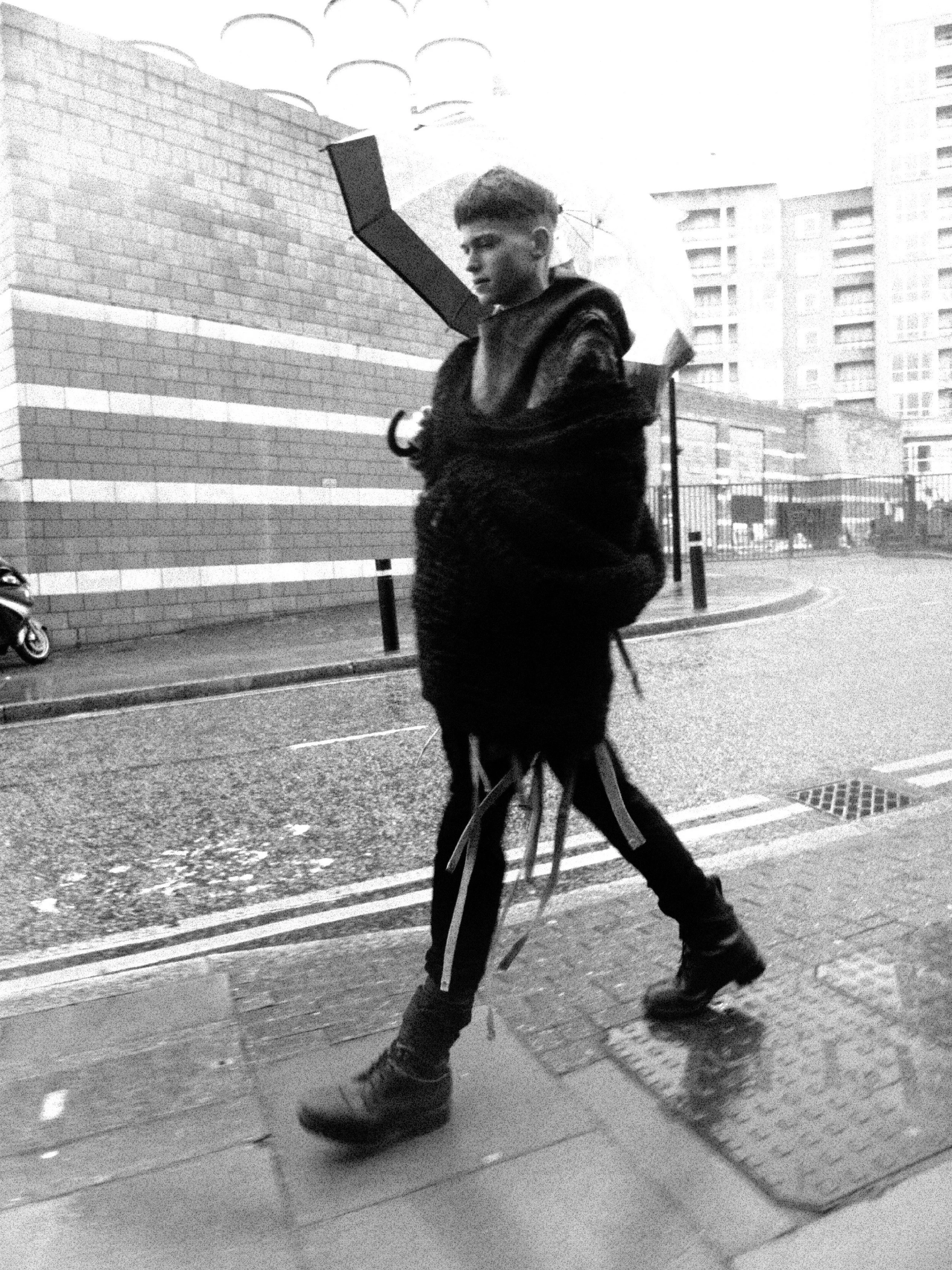A young person riding a bicycle on a city sidewalk, with a backpack and dressed in dark clothing, in front of modern buildings.