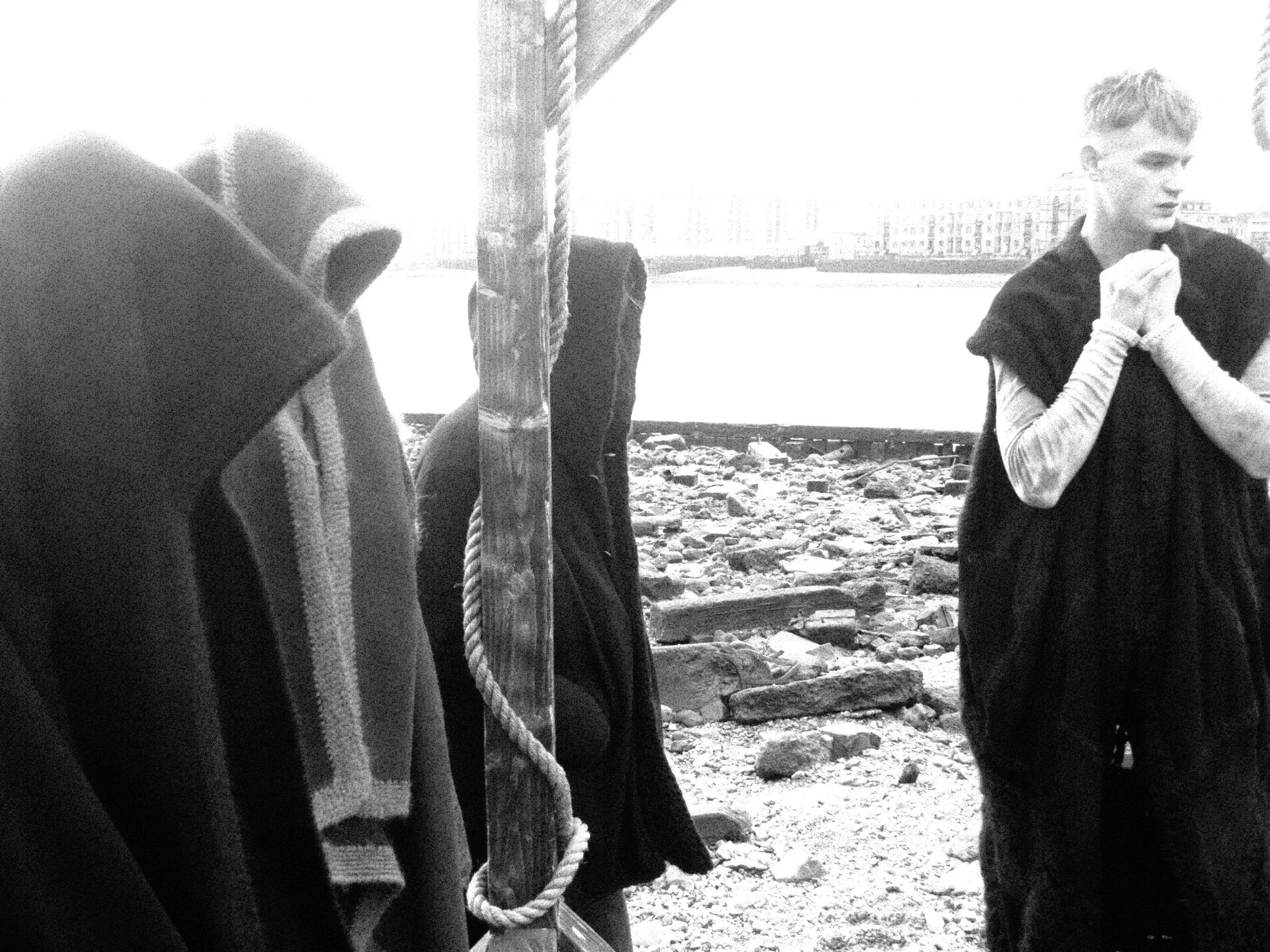 Group of people, including a young woman, on a beach near a body of water with buildings in the distance. Some individuals are partially obscured.