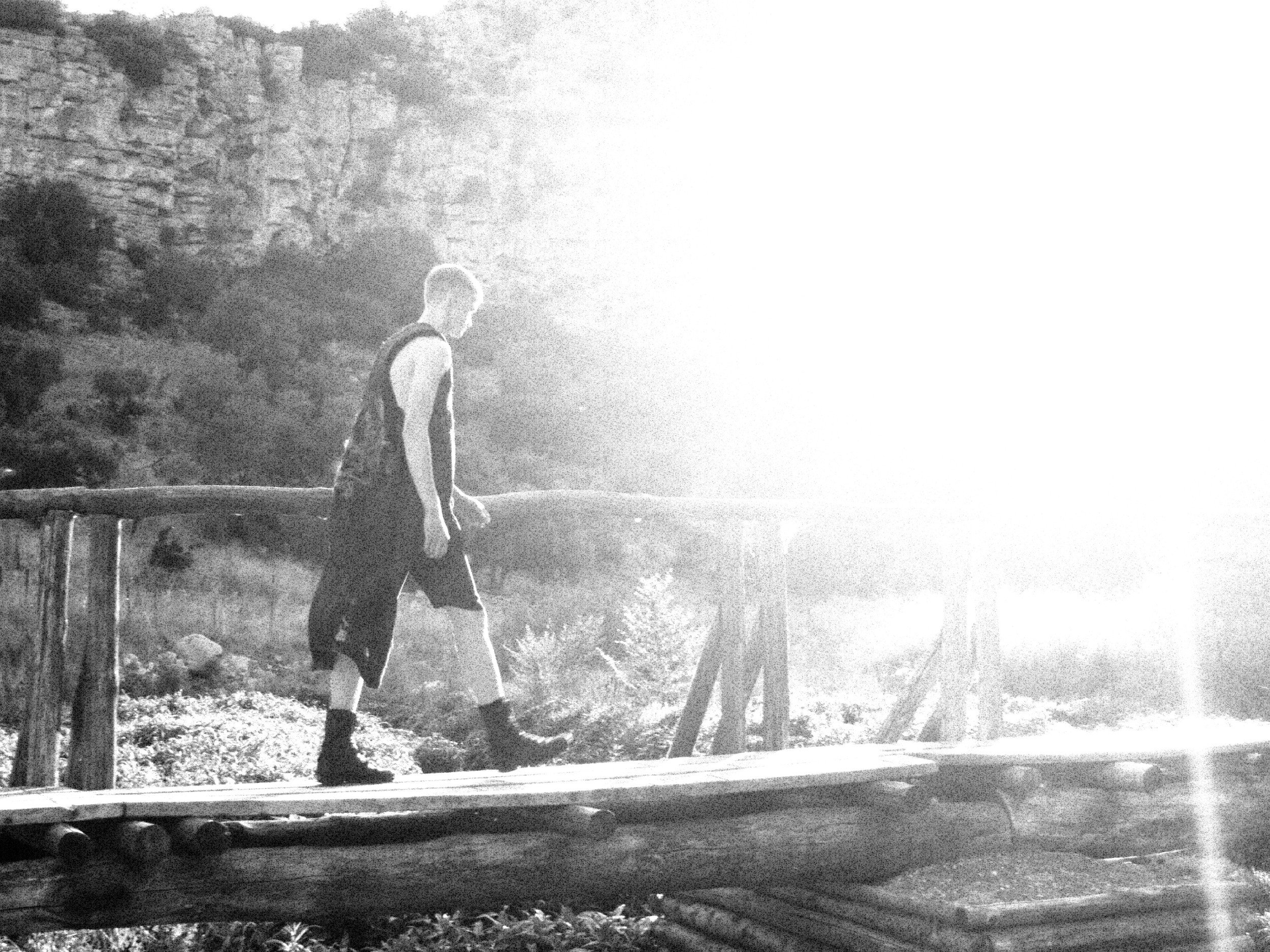 A person walking along a wooden bridge outdoors, with trees and rocky terrain in the background, in black and white.