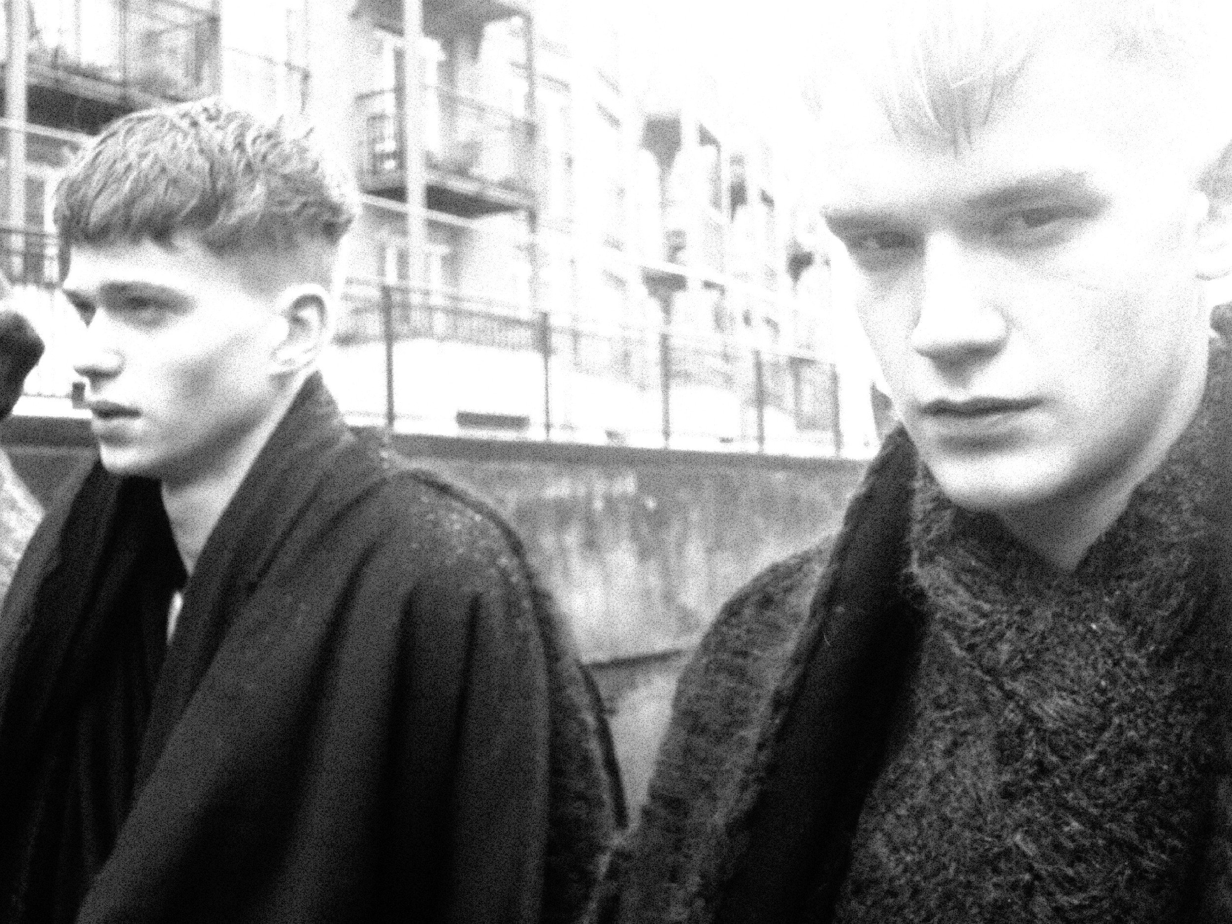 Black and white photo of two young men, one on the left with short wavy hair and the other on the right with slightly longer hair, standing outdoors in front of a multi-story building with balconies.
