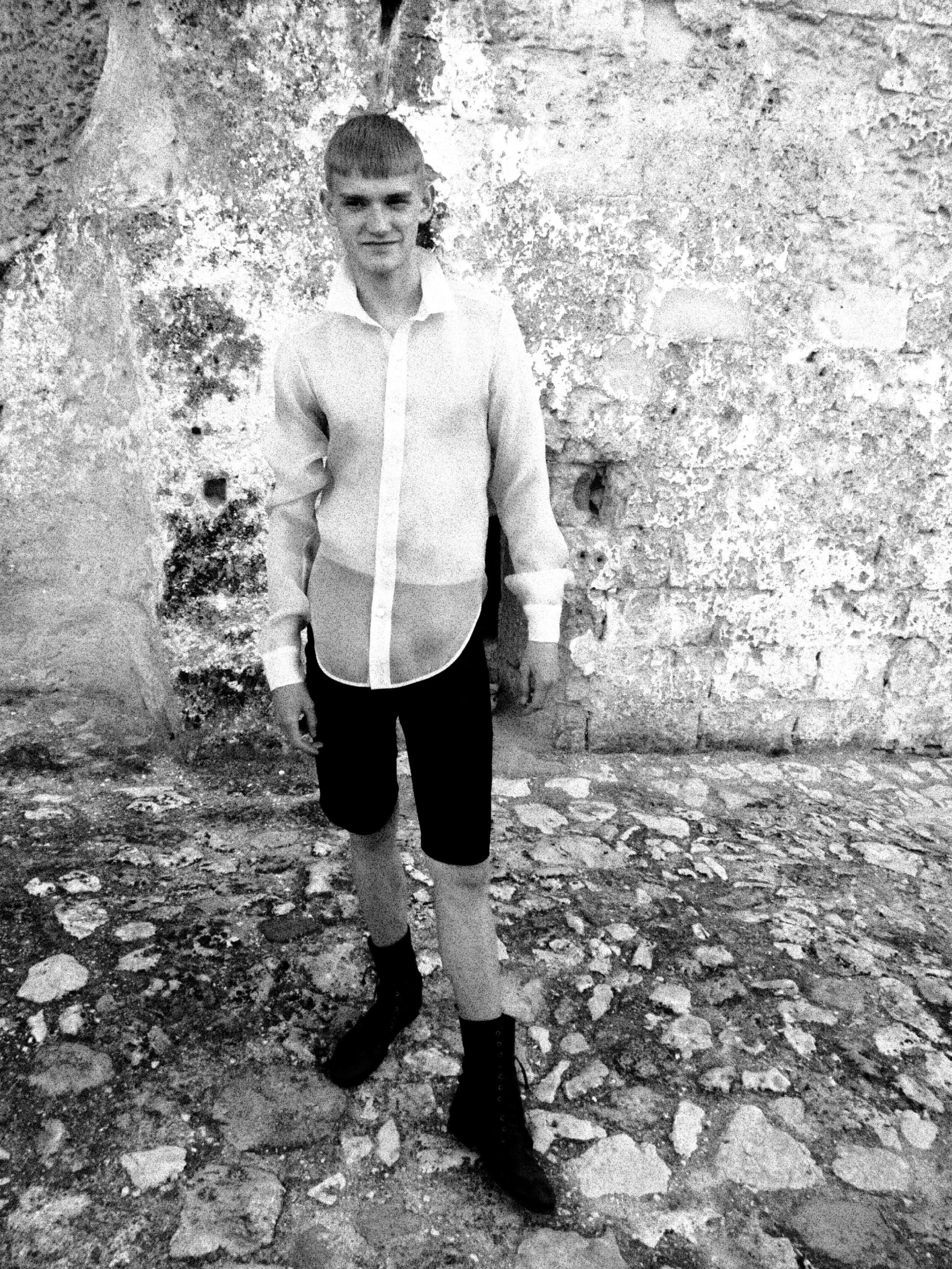 A person with short hair and a fringe, wearing a light-colored button-up shirt and dark pants, standing outdoors against a weathered stone wall. The ground is uneven with rocks and debris.
