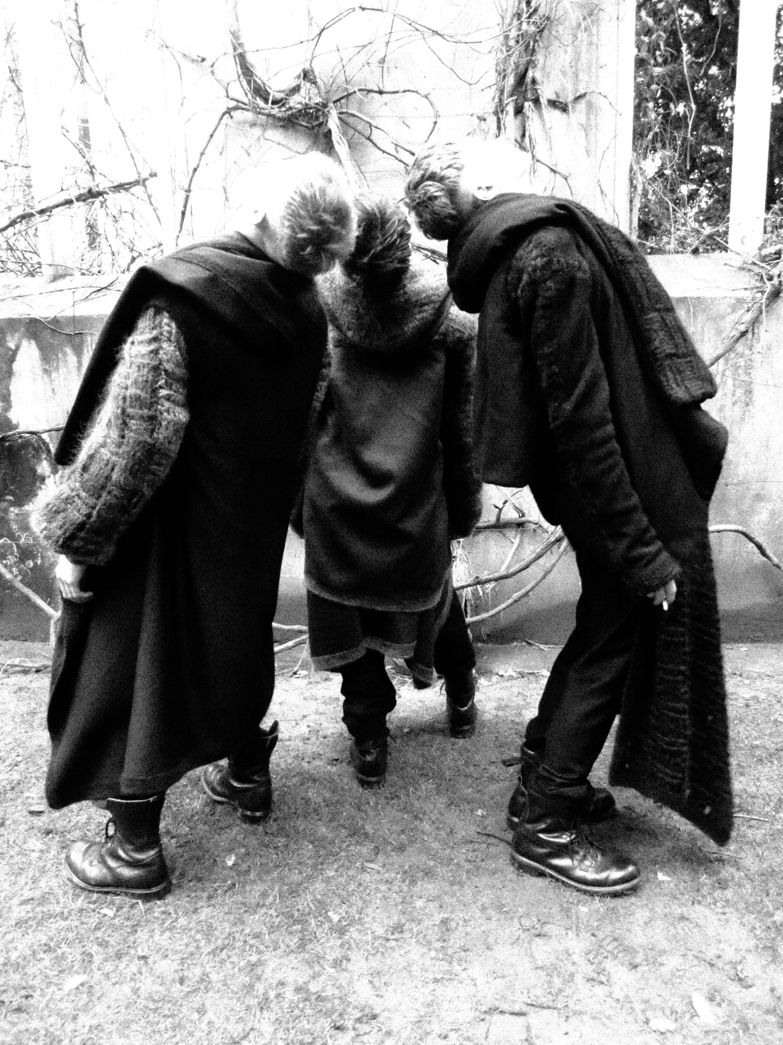 Four people are standing in a circle and looking down. They are wearing jackets, scarves, and boots. The background features a wall with tree branches and vines.
