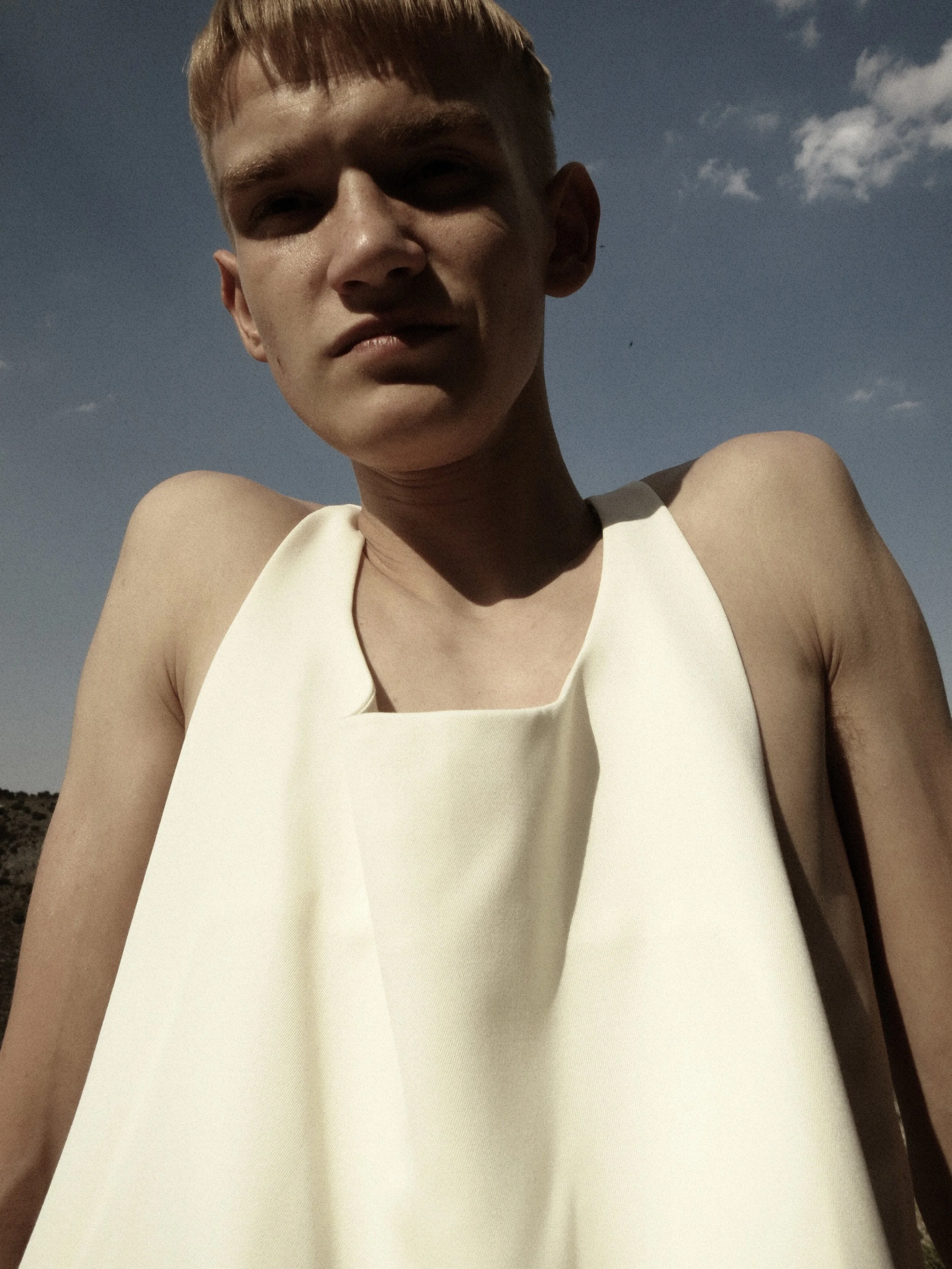 Close-up of a person with short blond hair wearing a sleeveless white top, standing outdoors against a blue sky with a few clouds.