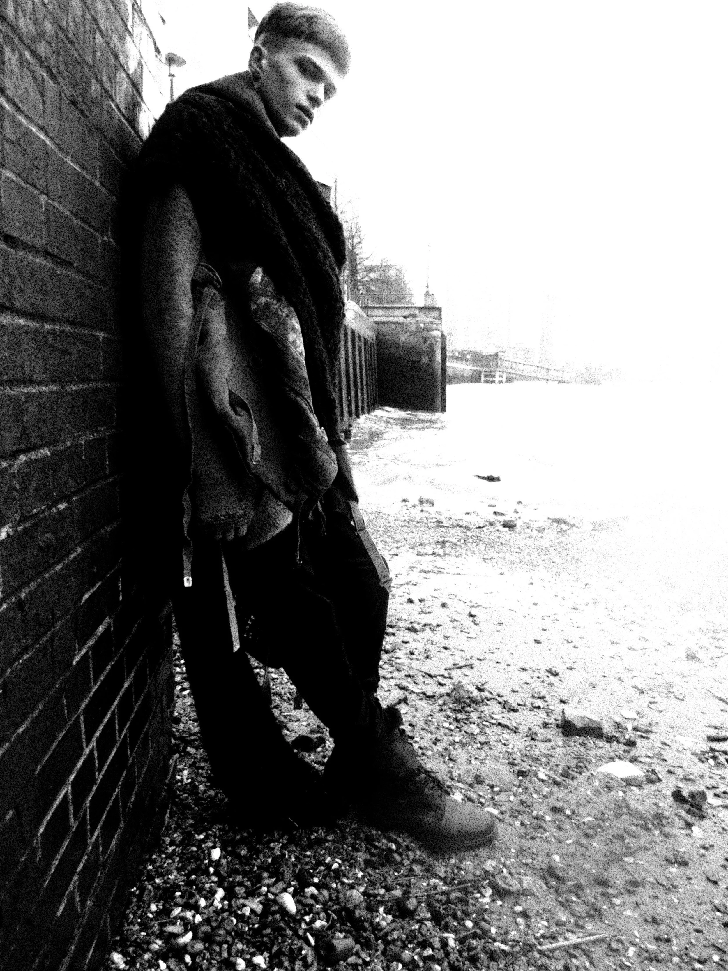 A young man leaning against a brick wall outdoors, looking down with a serious expression, in black and white.