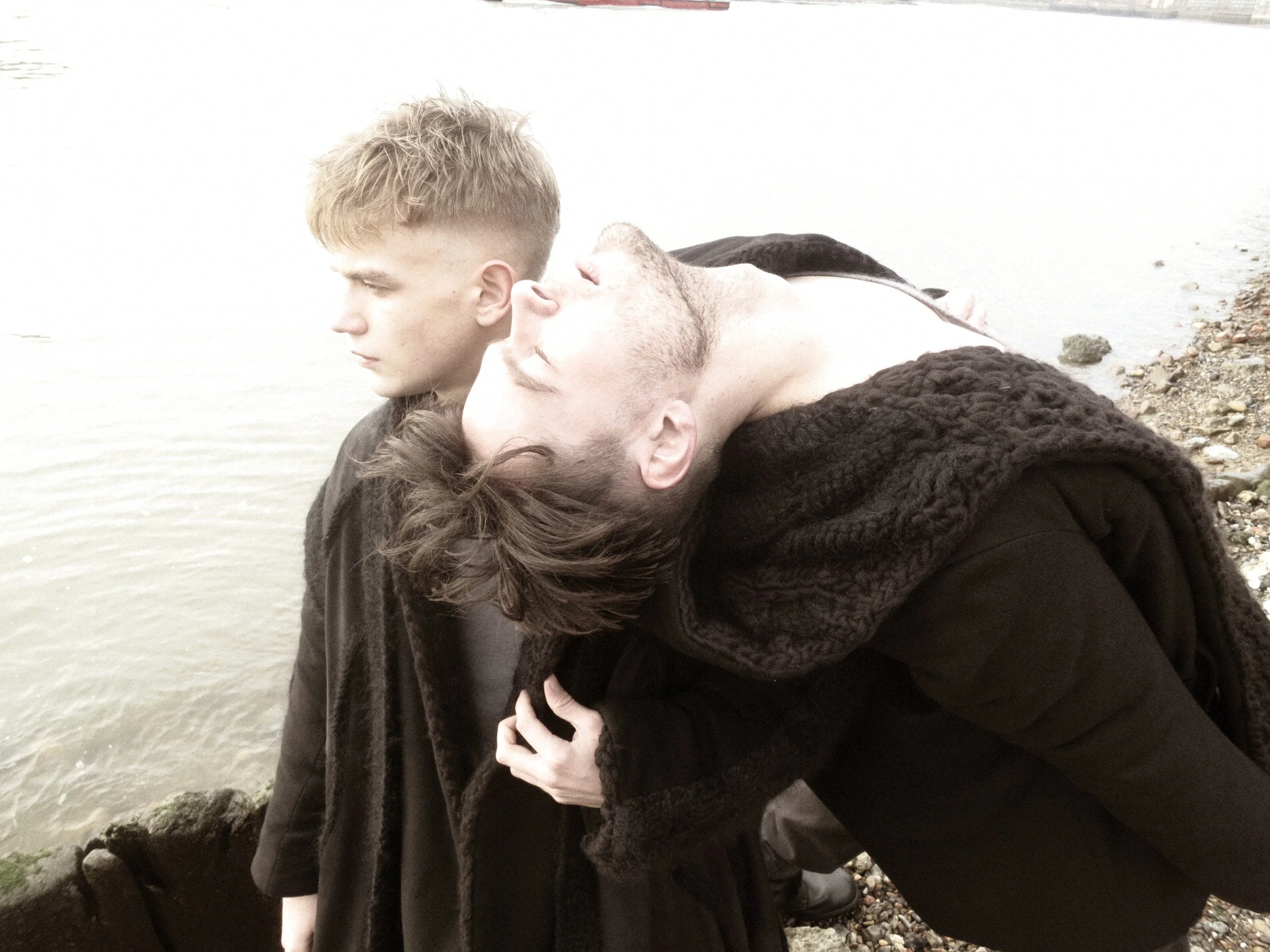 Two men are at the beach, leaning against rocks near the water, with one man appearing to kiss the other's head.