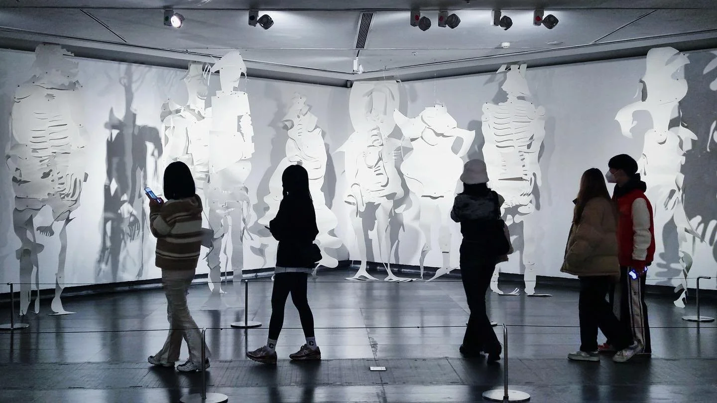 People viewing an art installation featuring white paper cutouts of human figures on a gray wall in an indoor museum setting.