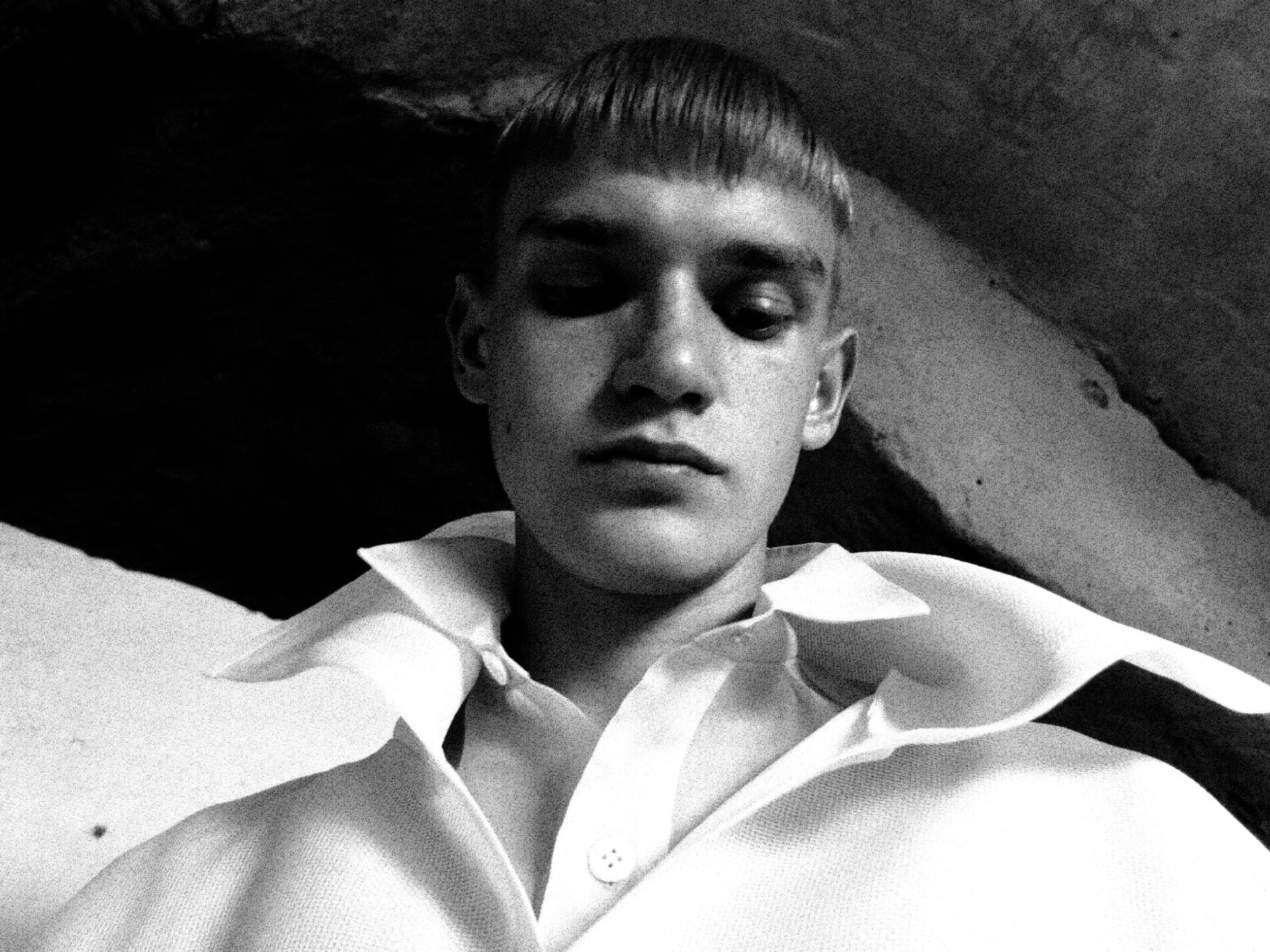 Black and white photo of a person's face with short hair, wearing a collared shirt, lying down on a soft surface.