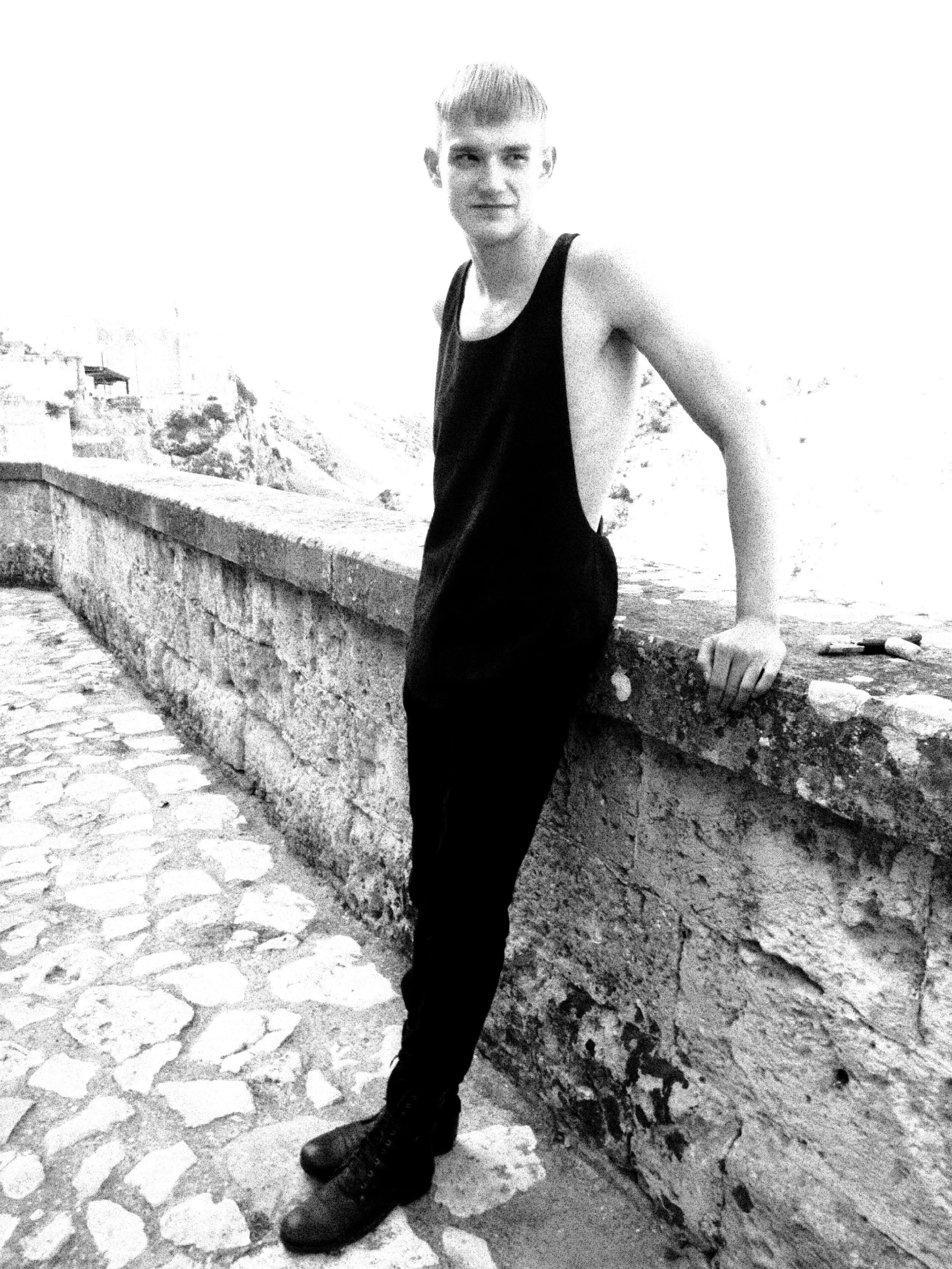 Black and white photo of a young man with a short haircut, wearing a sleeveless black shirt and black pants, leaning against a stone wall outdoors, with rocky terrain in the background.