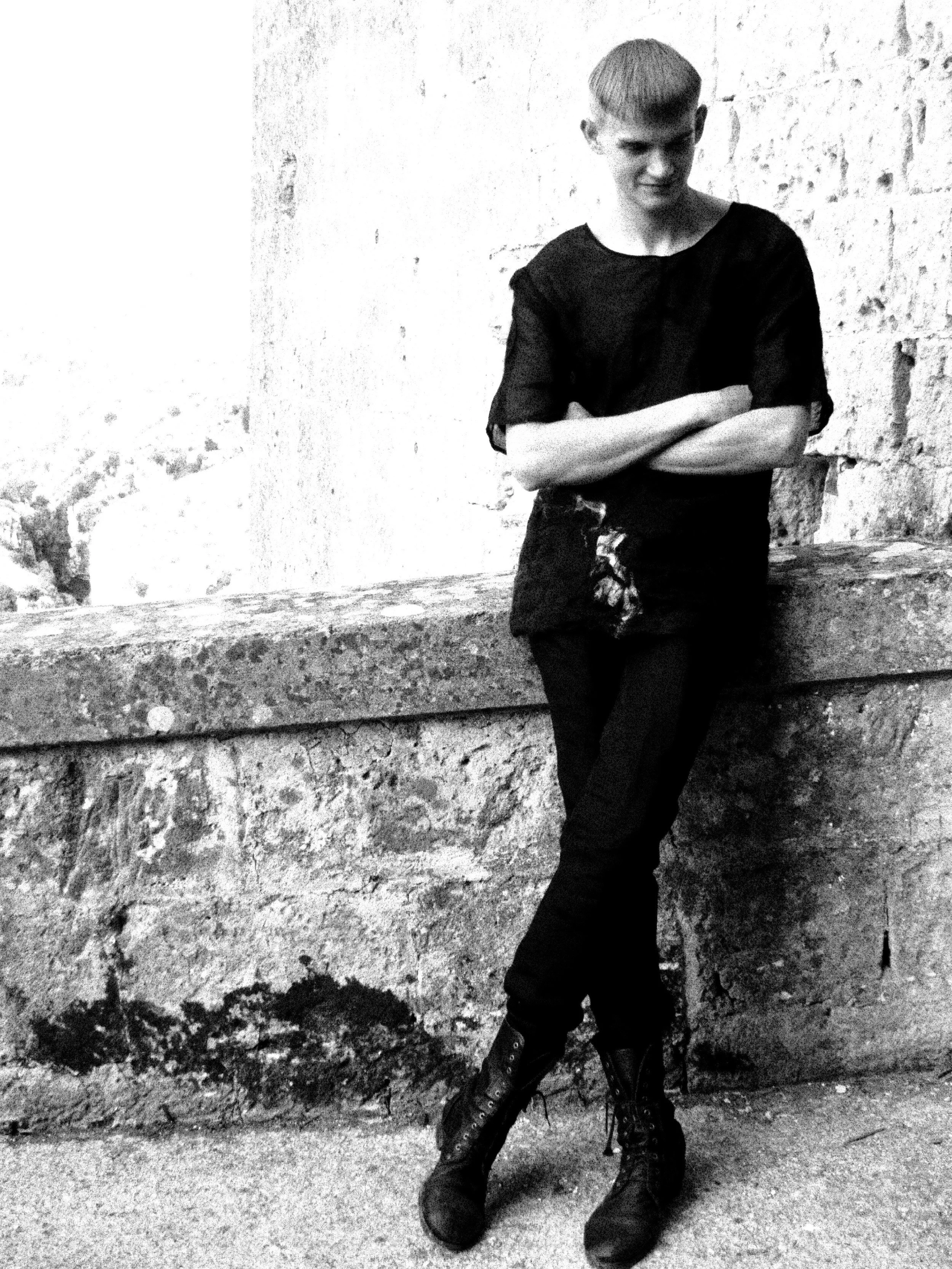 A person with a short haircut, wearing a black outfit and black boots, standing against a stone wall with arms crossed.