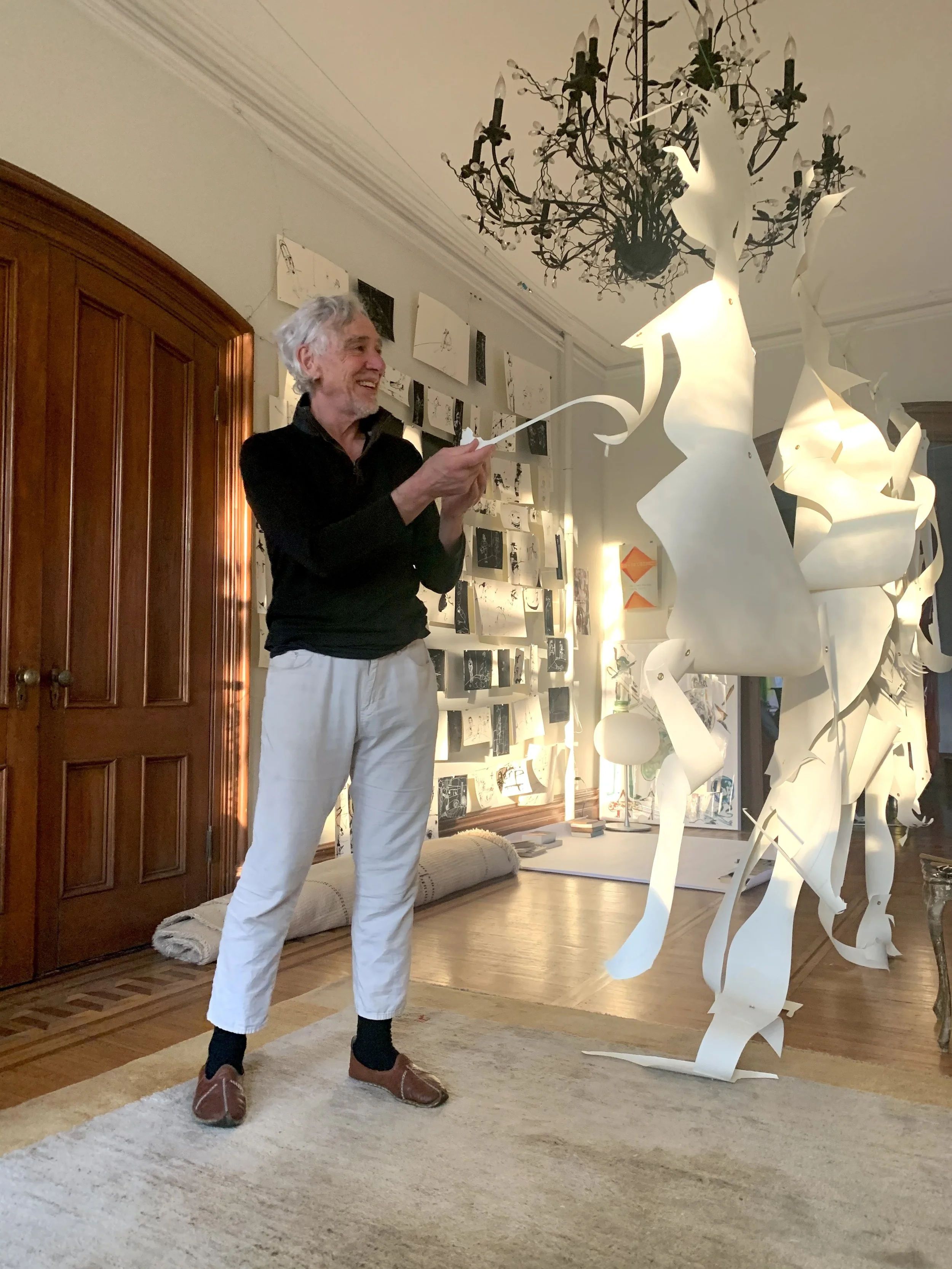 An artist is working on a paper sculpture in a brightly lit art studio with a chandelier. The artist is smiling and holding a piece of paper. The studio has walls decorated with sketches and drawings.