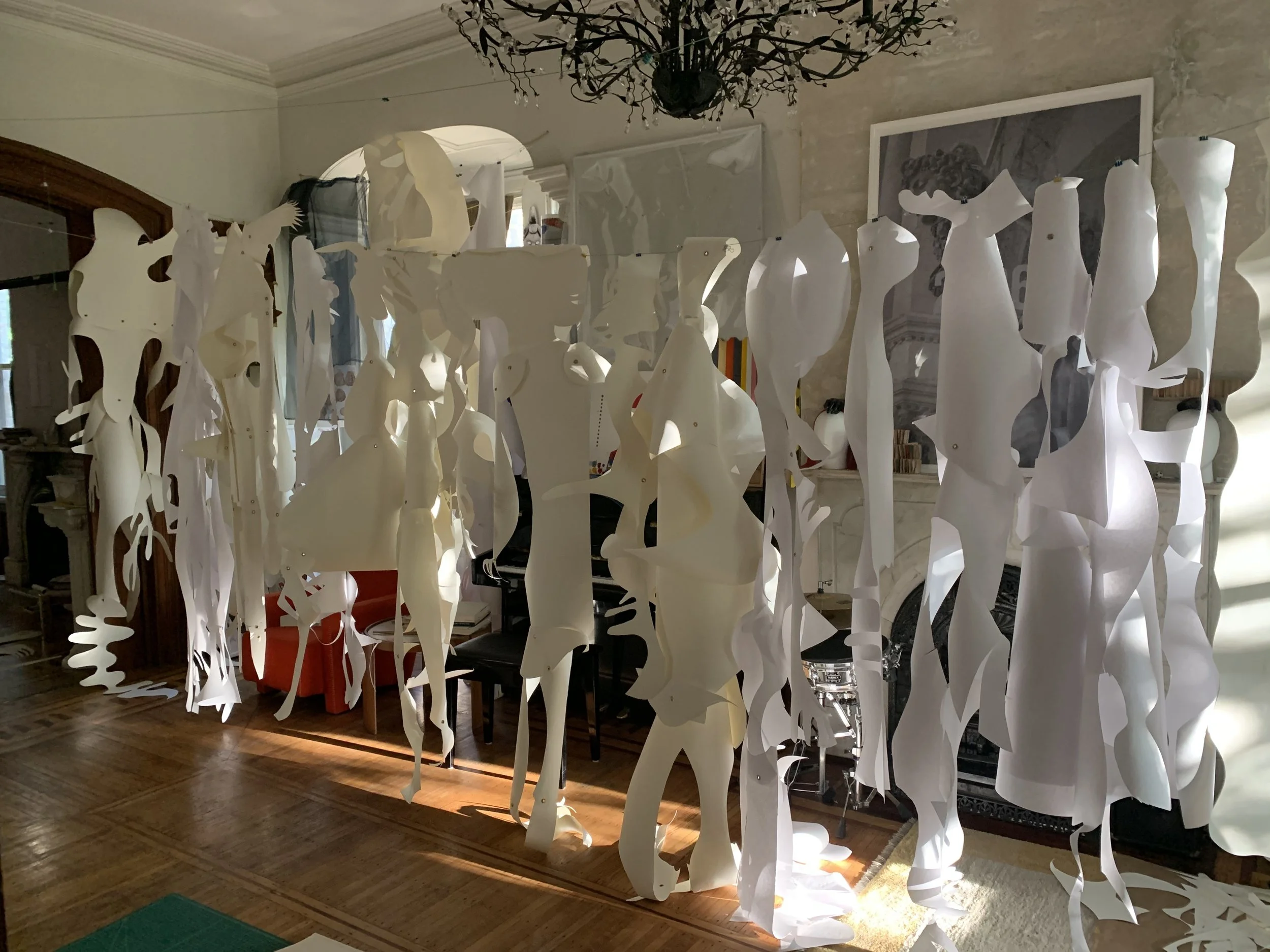 Indoor art installation featuring various white abstract paper sculptures hanging from the ceiling inside a room with wooden floors and a fireplace.
