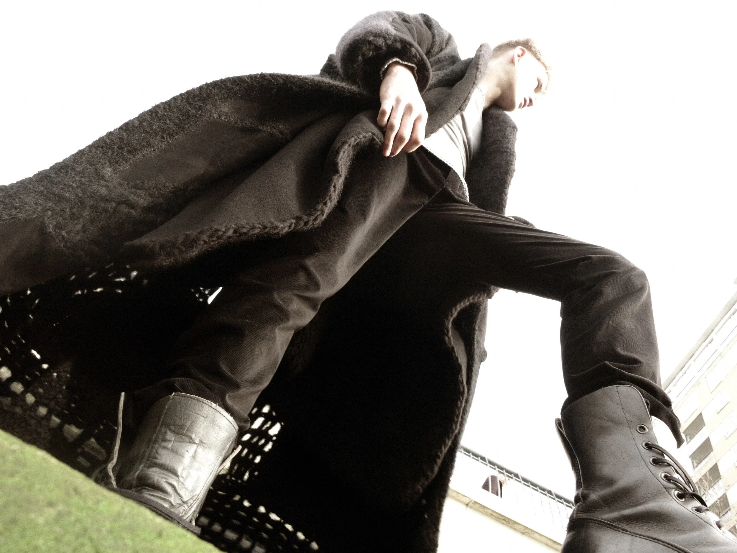 A person wearing black boots, black pants, a long dark coat, and a hoodie, sitting on a wire chair, with an indoor background.
