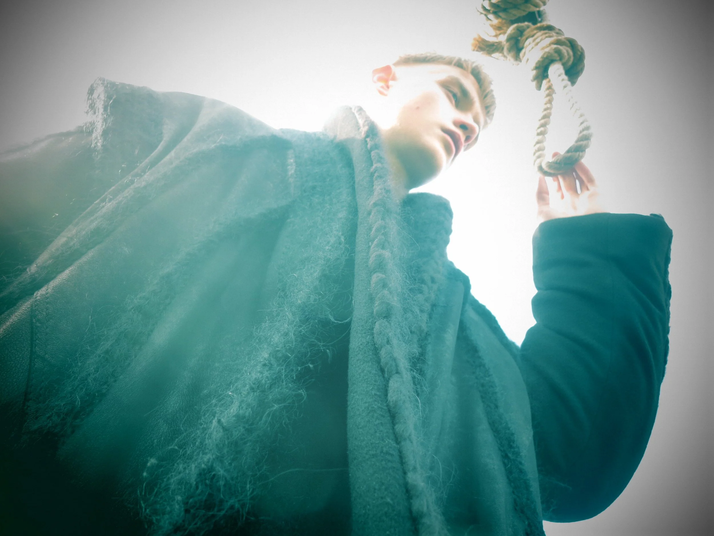 A person with blonde hair holding a noose hanging from above, wearing a dark hoodie, looking up toward the noose.