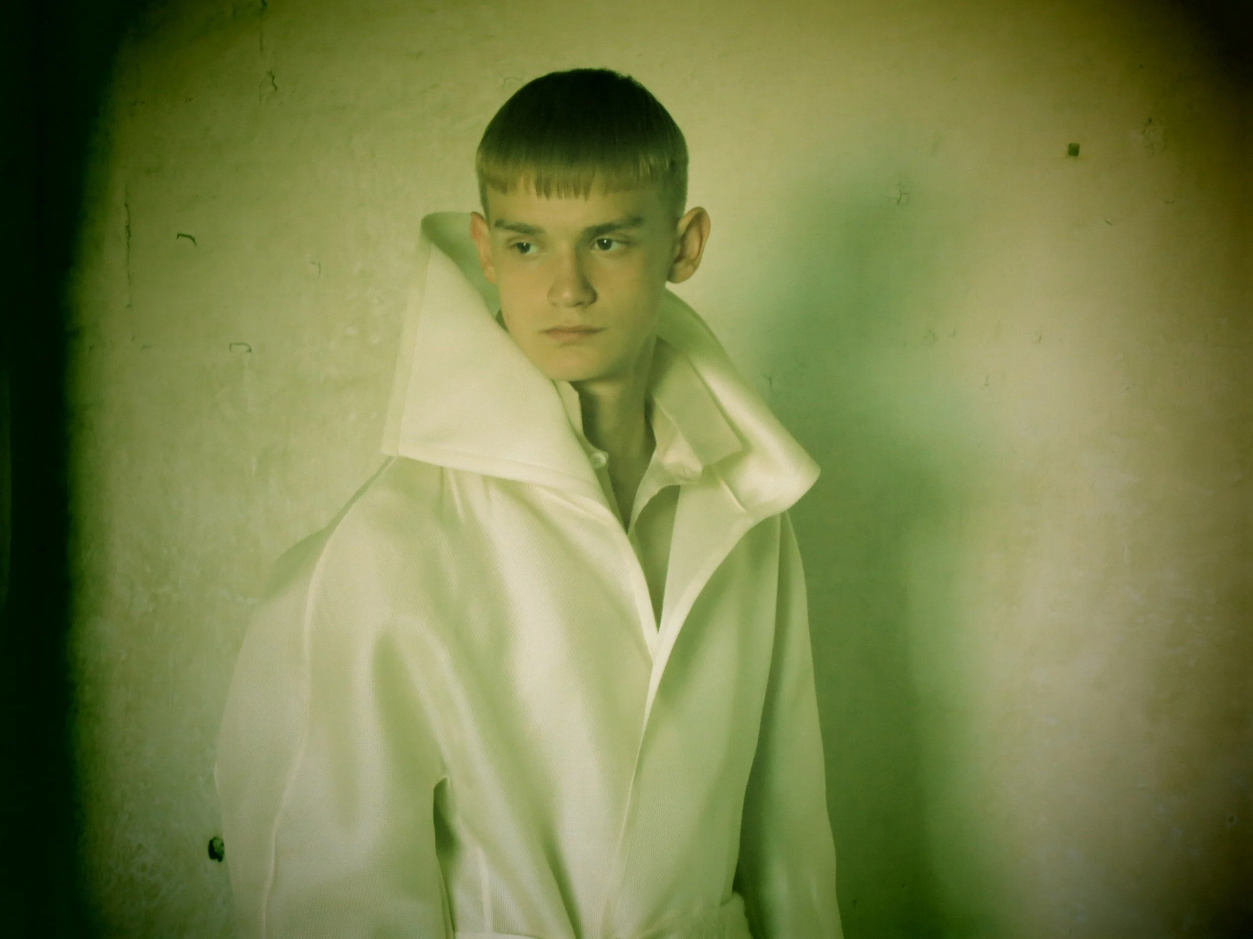 A young boy with short dark hair and a serious expression wearing a white shirt, leaning against a beige wall with a greenish tint.