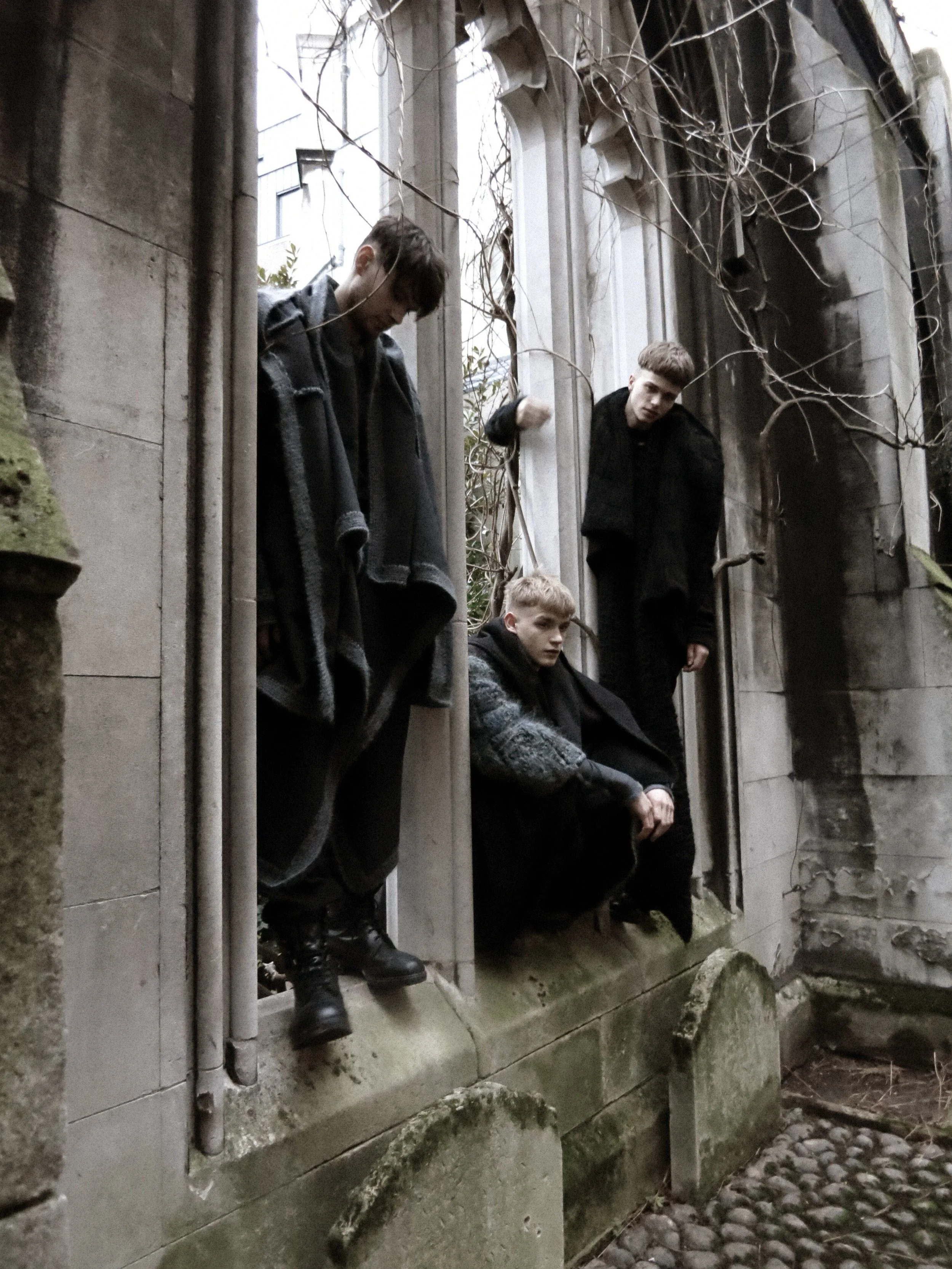 Four young men with dark clothing and messy hair are standing and sitting on the ledge of a stone building with gothic architecture, overgrown with vines and leaves, in what appears to be an urban or historical setting with cobblestone pavement nearb