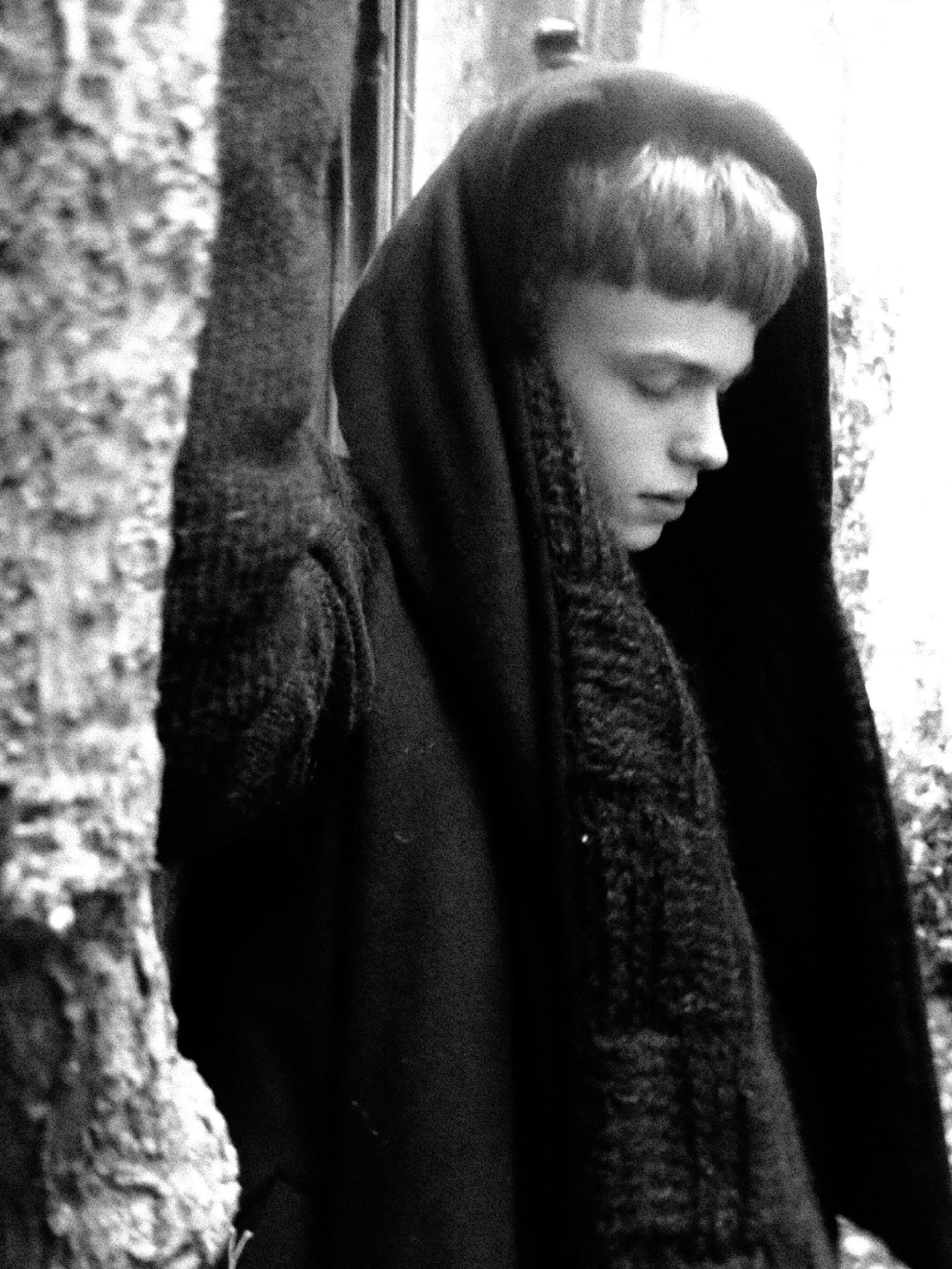 A young woman with short hair and bangs, wearing a hooded jacket, looking downward, standing near a textured wall.