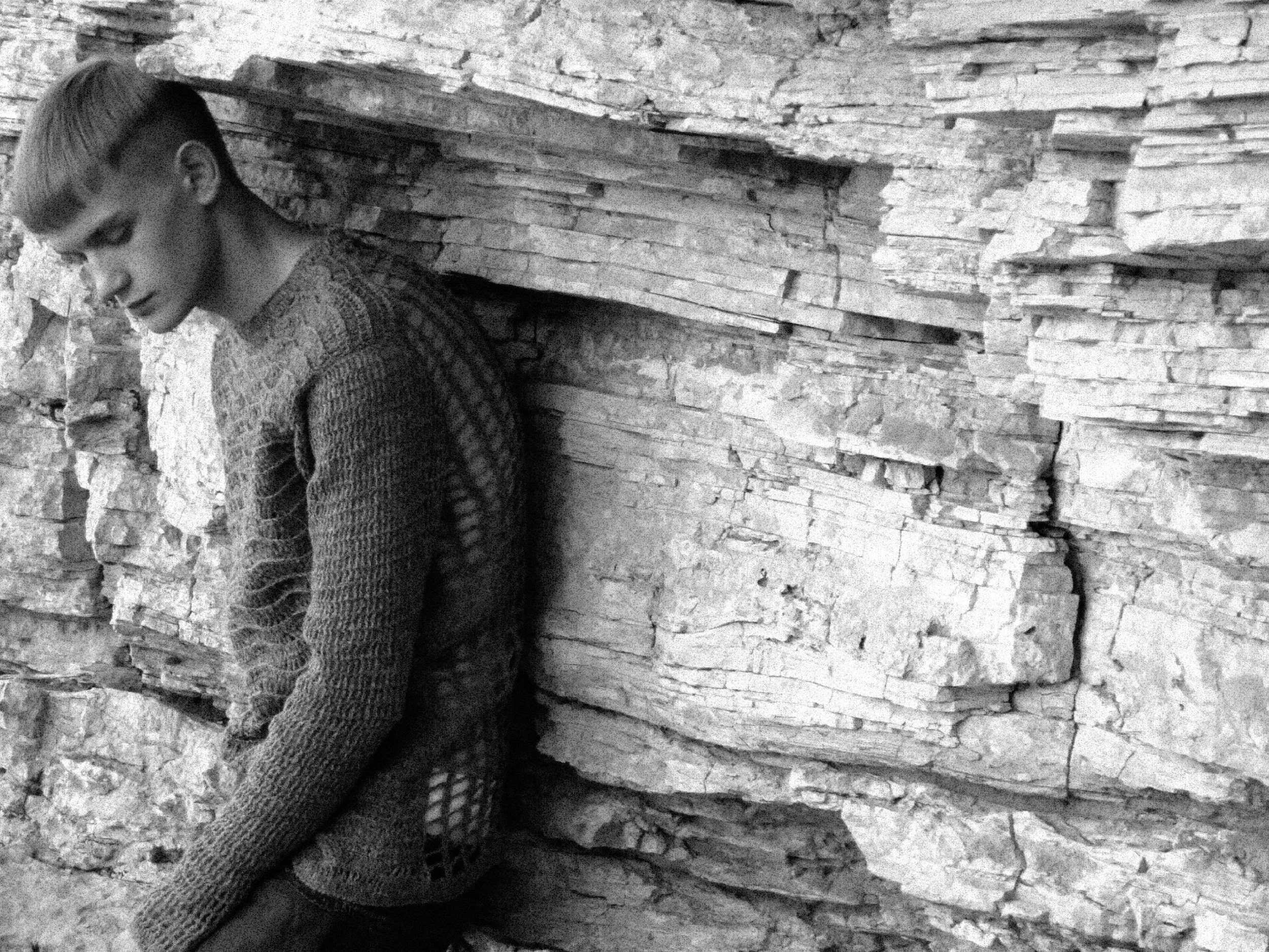 A person with a bowl haircut and patterned sweater leaning against a textured stone wall.