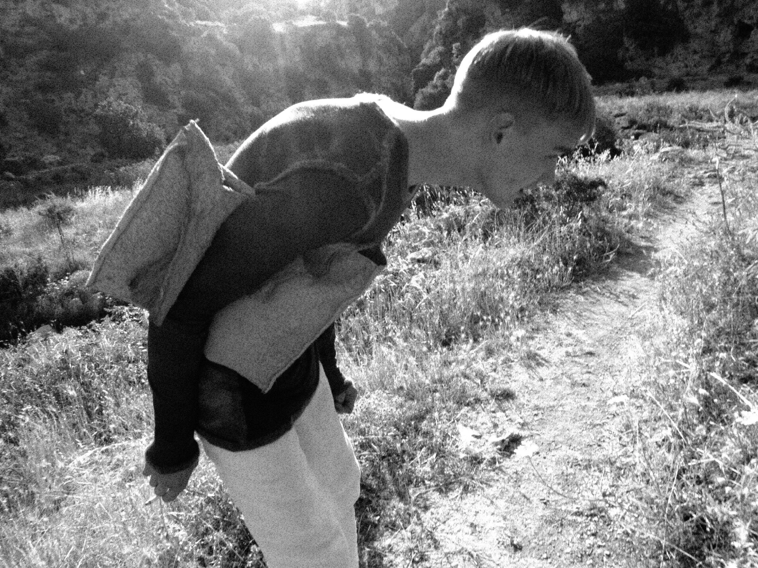 A person stands outdoors in a scenic, natural landscape with hills and trees, captured in black and white. The individual is facing away, holding a child's hand, and appears to be looking down or toward the ground.