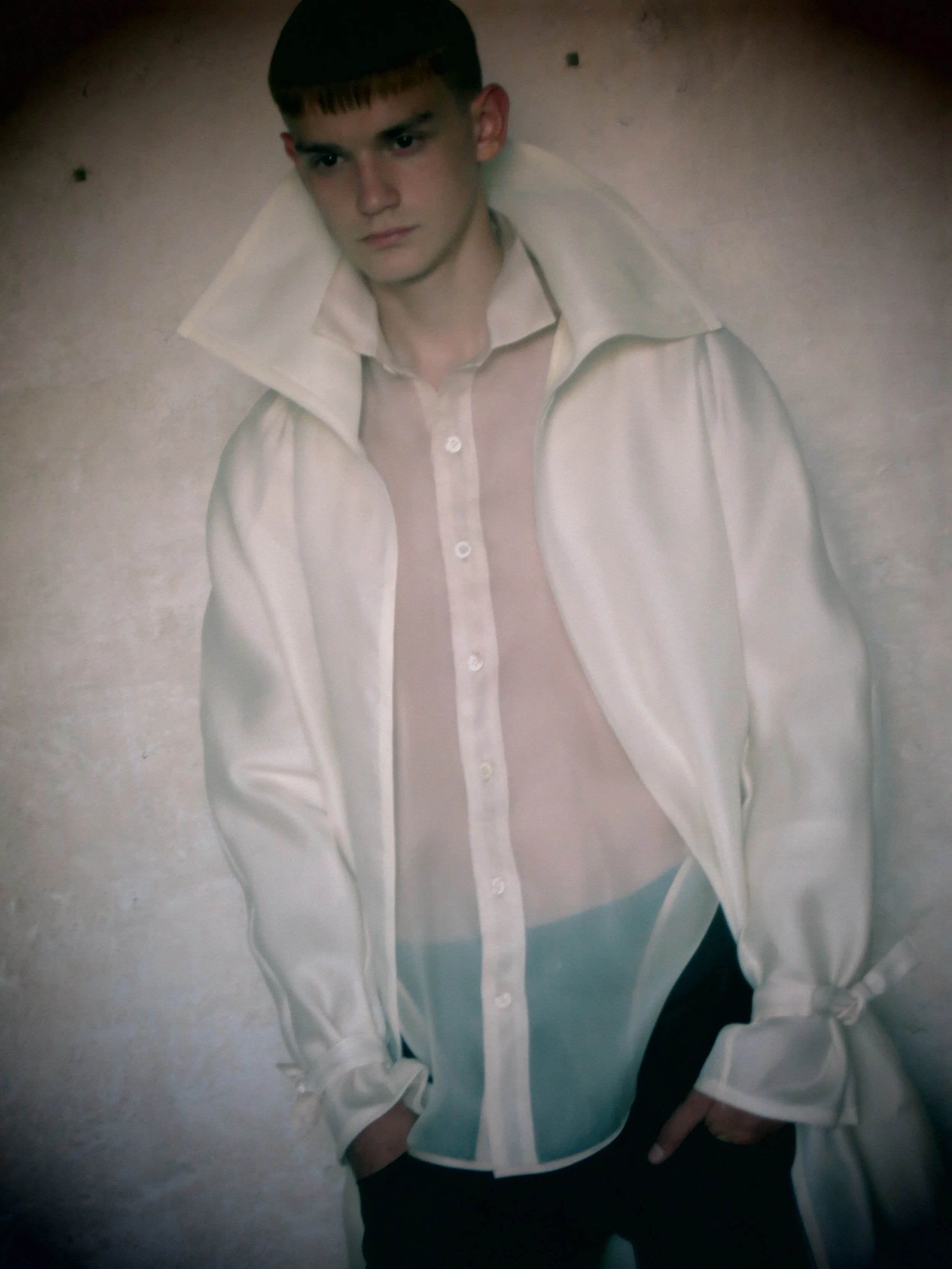 A young man wearing a white button-up shirt with a collar and black pants, standing against a plain wall with a serious expression, his hands in his pockets.