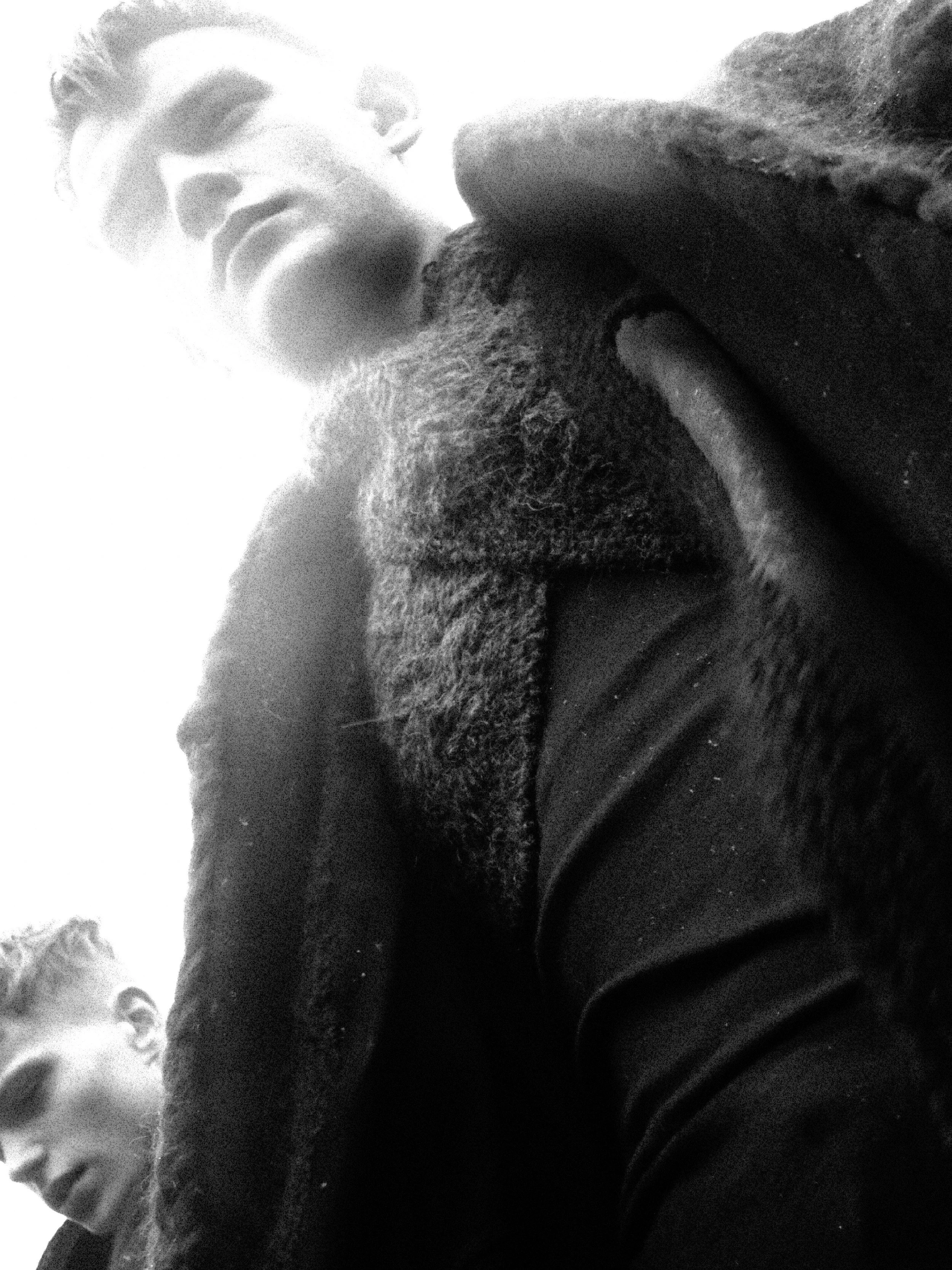A black and white close-up photo of a young man, looking upwards with his mouth open, wearing a black shirt, with short curly hair and a beard.