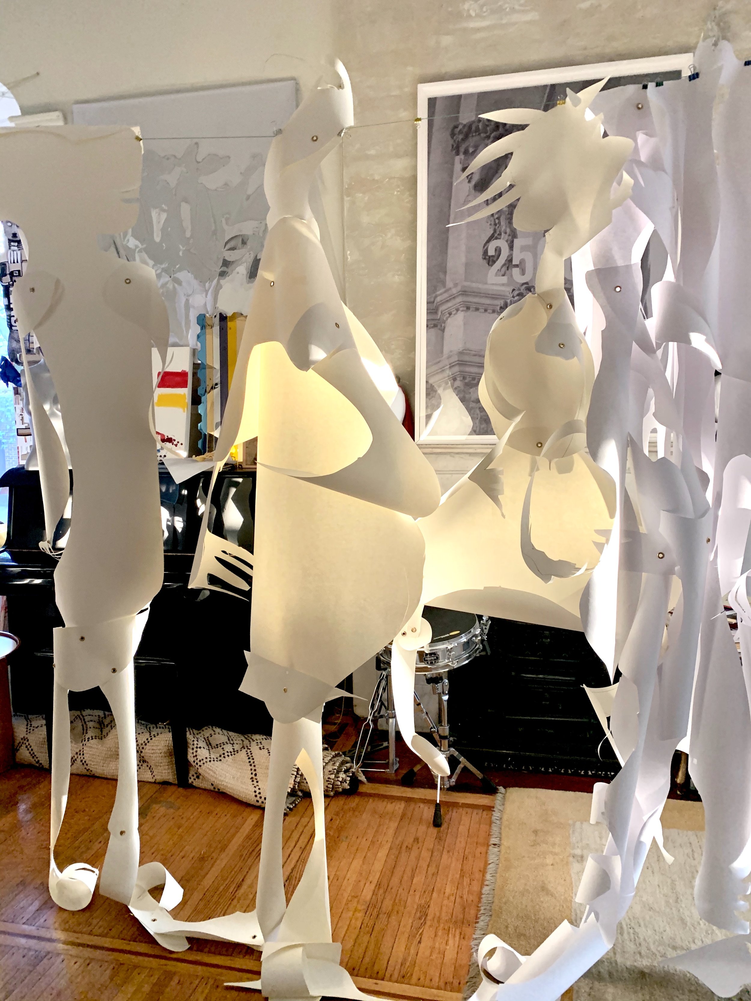 Art installation featuring large white paper or fabric sculptures suspended in a room with a wooden floor, with musical instruments such as a drum set and a piano, and framed artwork on the wall in the background.