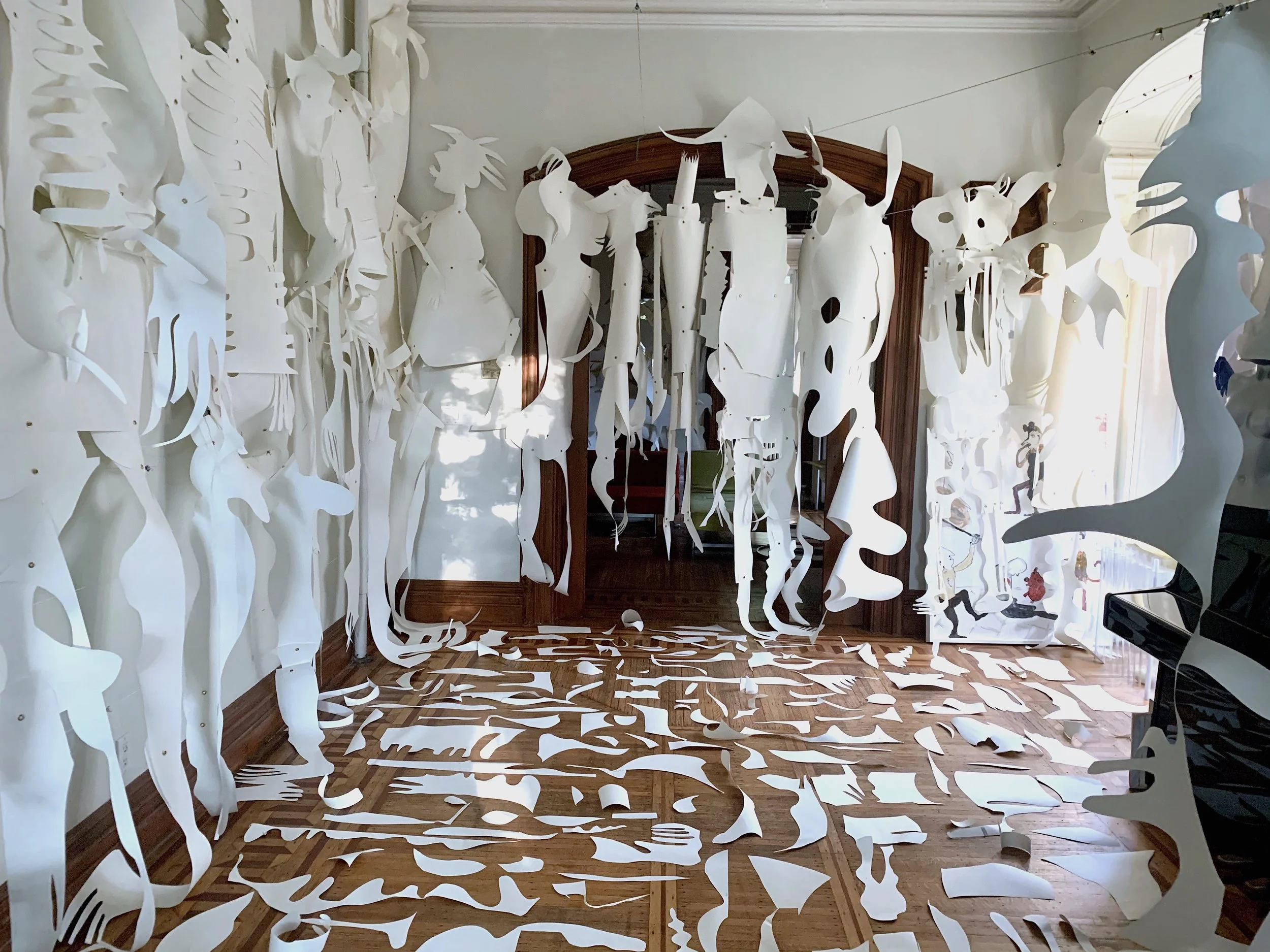 Room decorated with numerous white paper cutouts of abstract and organic shapes hanging from the ceiling and scattered on the wooden floor, with a large mirror and a grand piano visible.