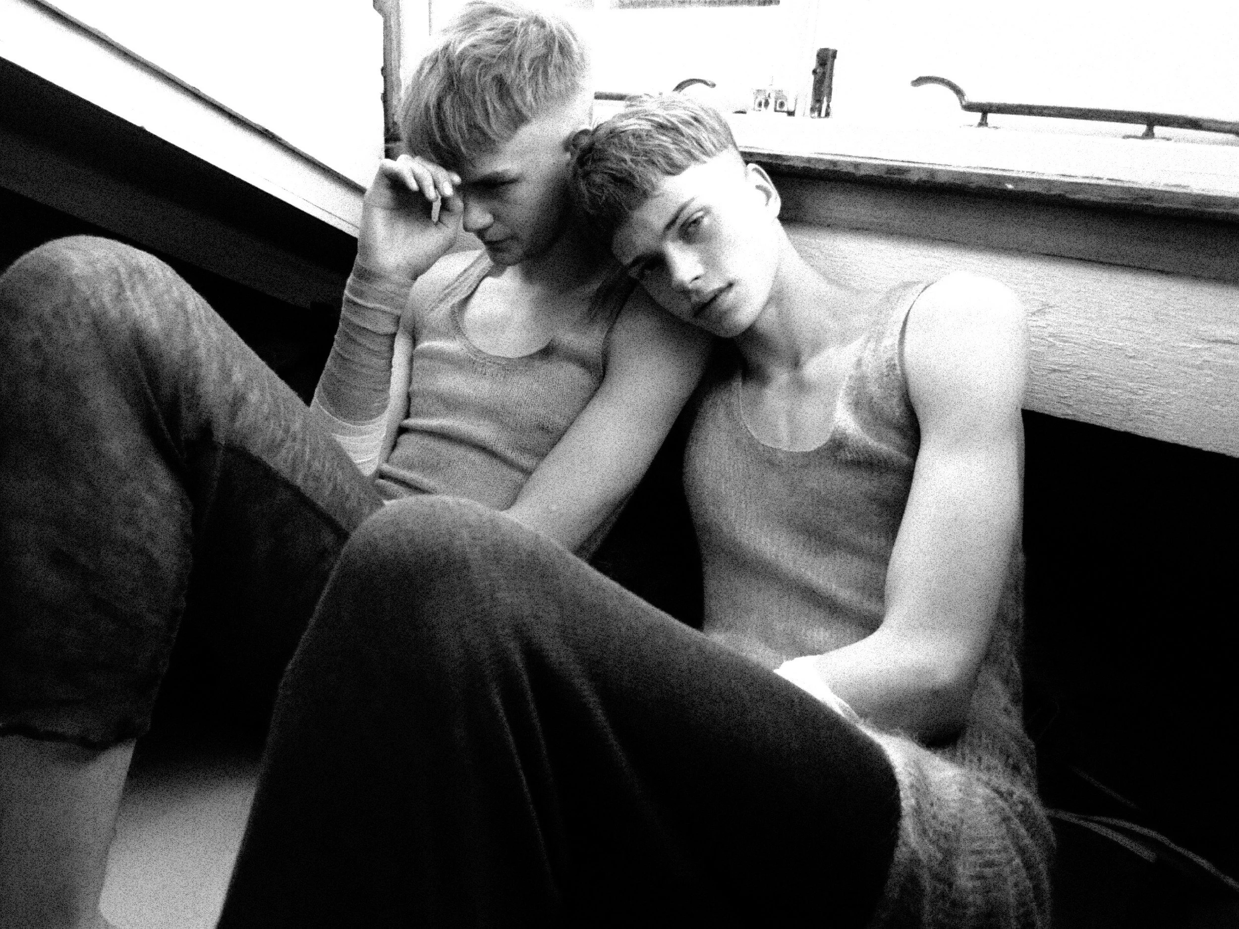 Two young men sitting close together on a bench, one with his head resting on the other's shoulder, in a black and white photo.