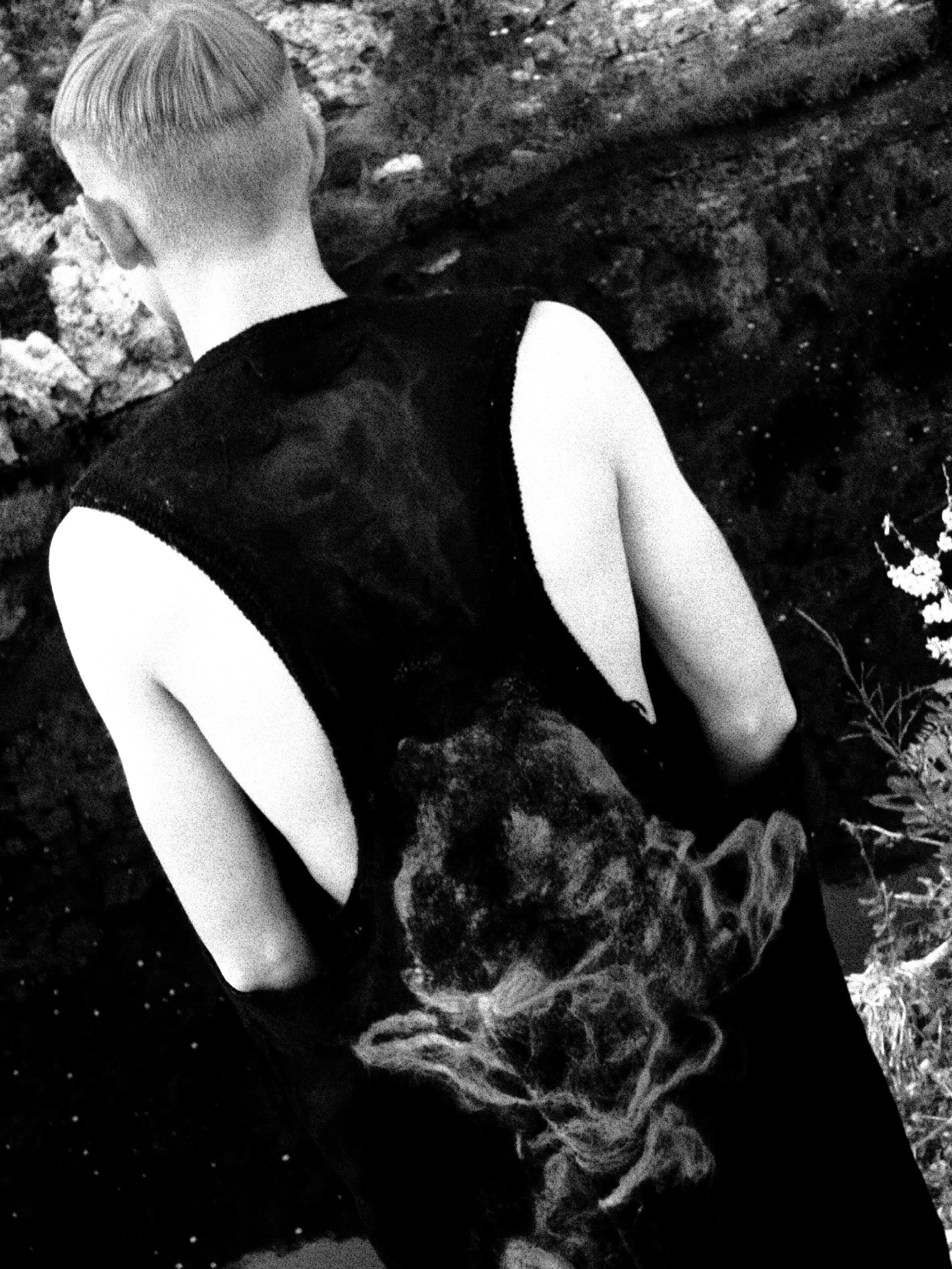 A person with short hair, seen from behind, standing in front of a textured rock or stone surface surrounded by foliage.