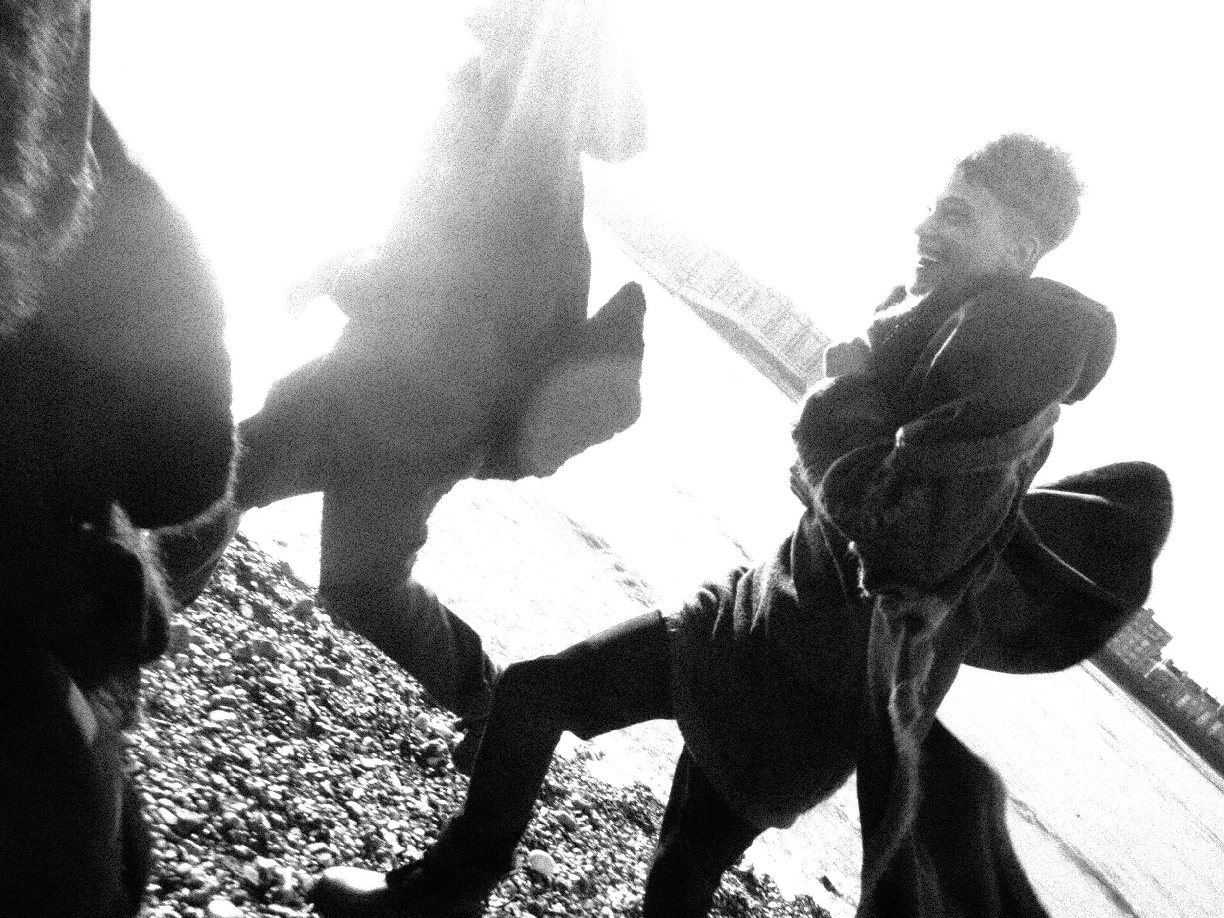 Black and white photo of three children playing outside. One girl with curly hair is on the left, another girl with short, curly hair and a jacket is on the right holding a guitar, and a boy with short hair is in the center holding a book or tablet. 