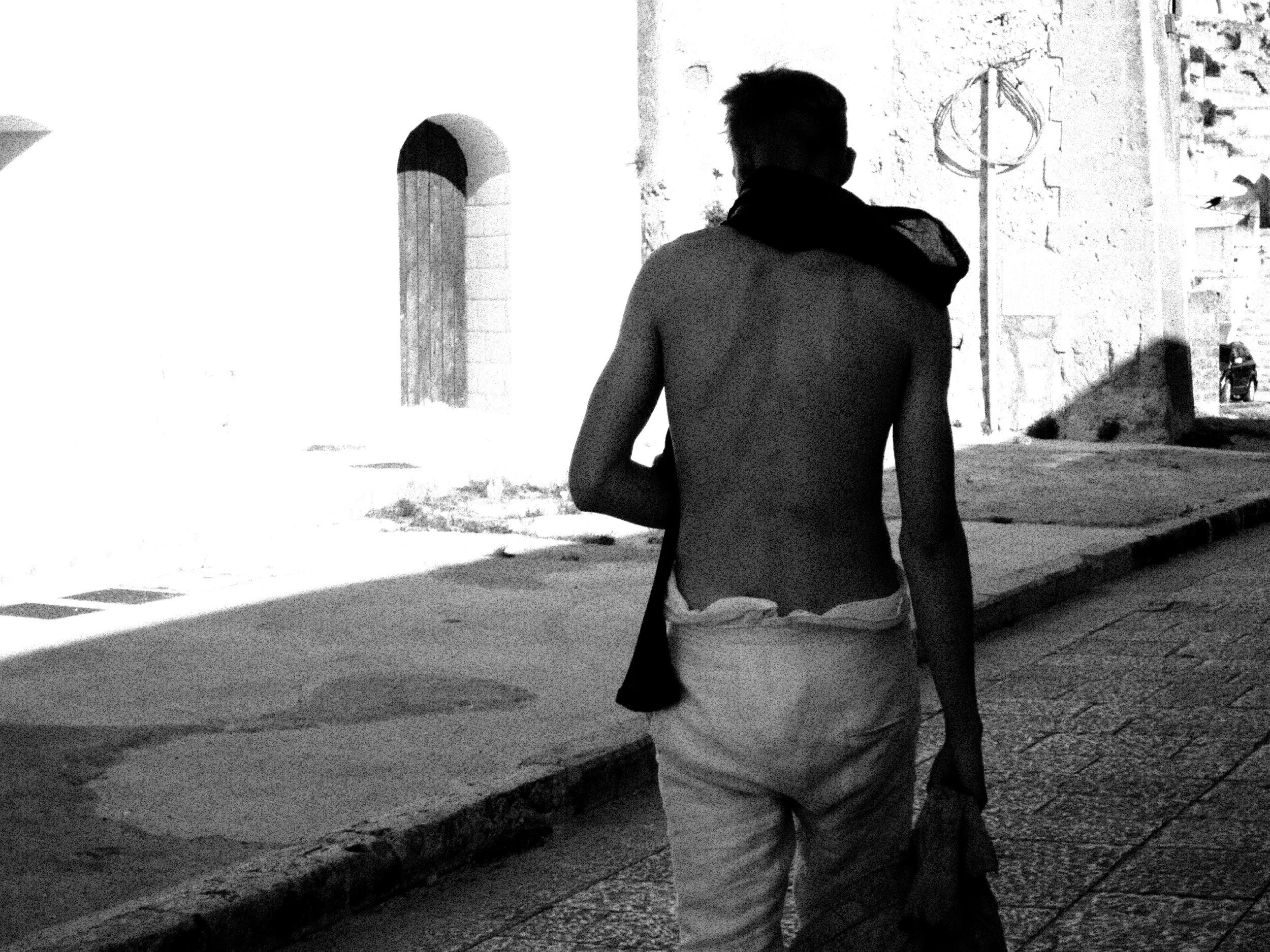 A shirtless person with short hair walking down a city street at night, with old stone buildings and a bicycle in the background.