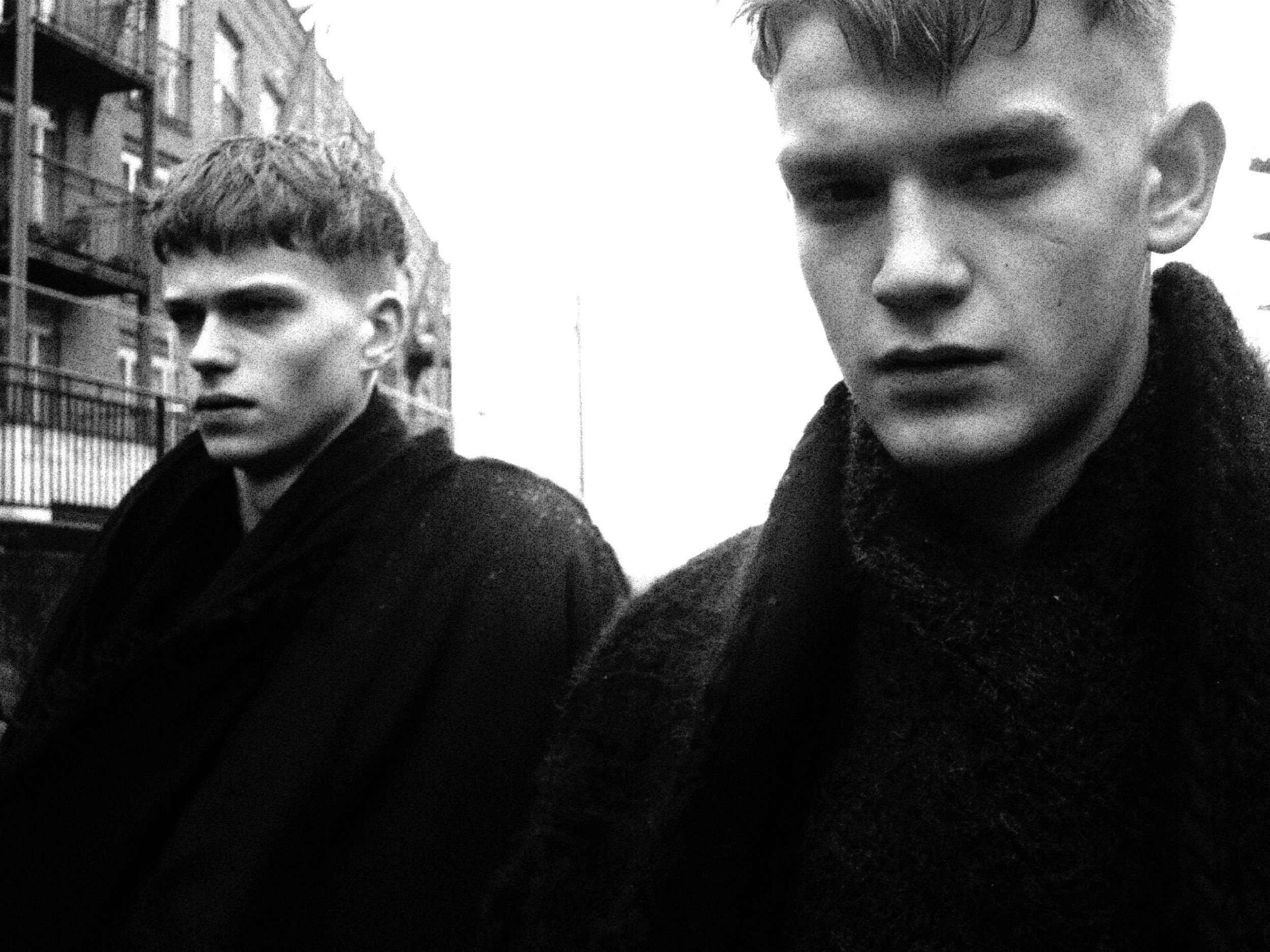 Black and white photo of two young men standing outdoors, near apartment buildings, with serious expressions.
