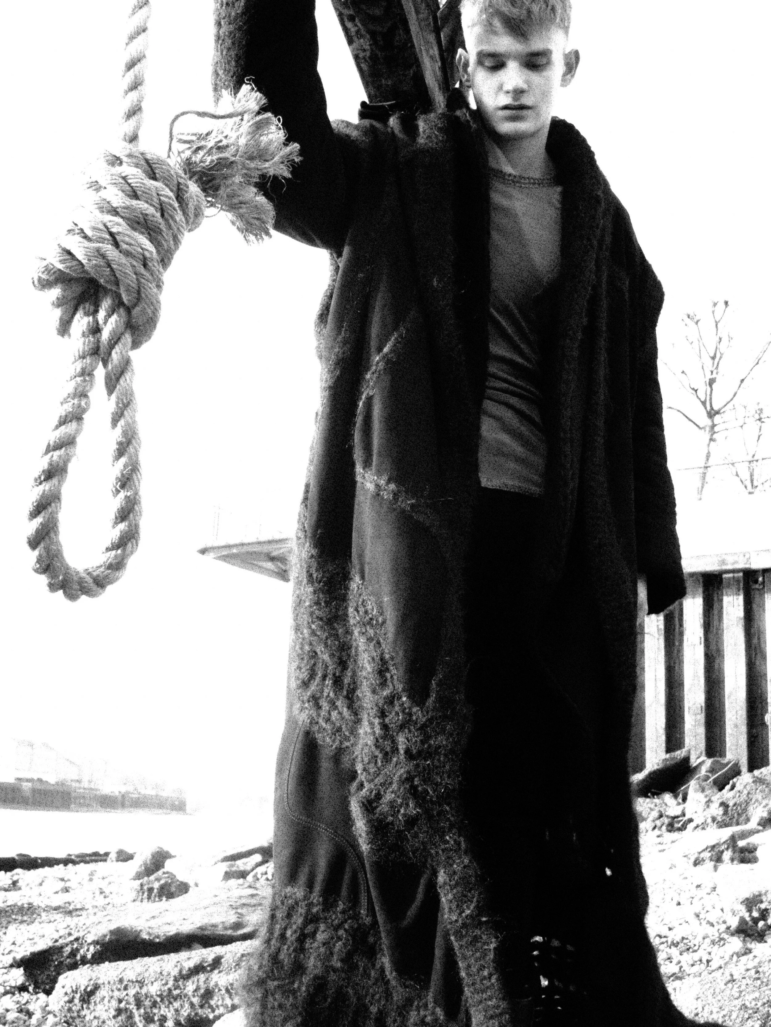 A black and white photo of a young person, possibly a male, with short hair, leaning against a large piece of wood or tree trunk. They are outdoors, with a fence and leafless trees in the background. The person wears a dark coat or jacket, and they a
