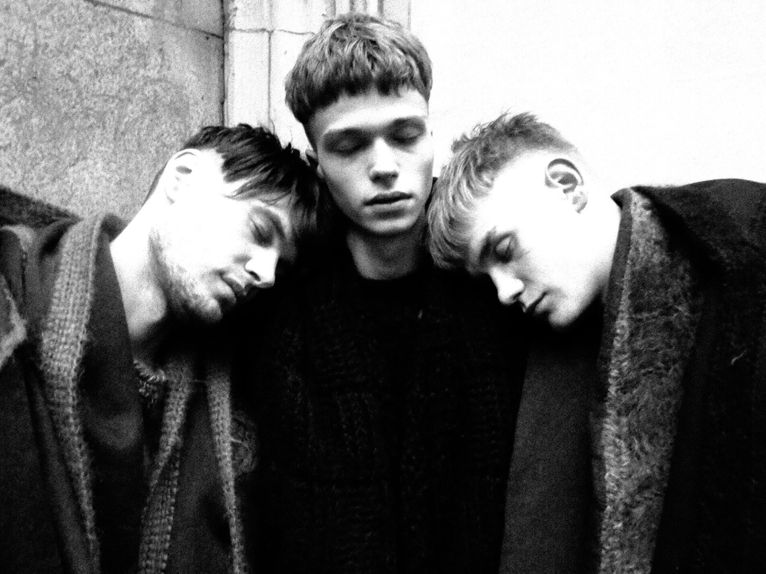 Black and white photograph of four young people, with three of them resting their heads on each other's shoulders close together, eyes closed, and appearing peaceful or asleep. They are sitting outdoors near a stone wall.