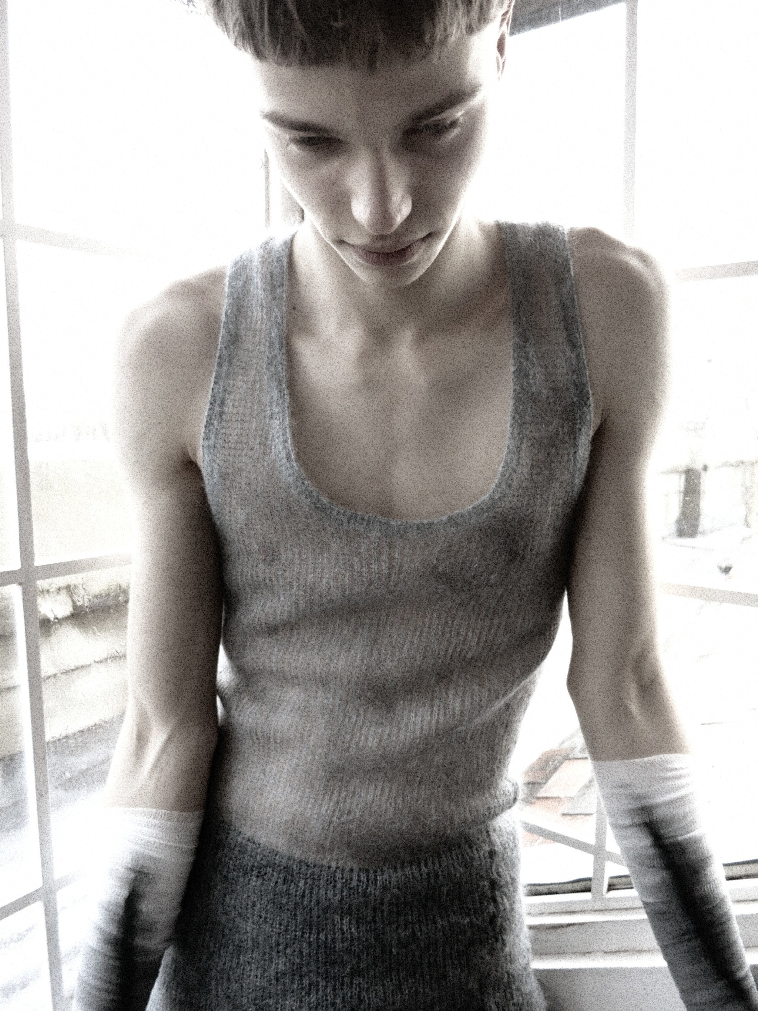 Young person, wearing a sleeveless top and long glove, leaning on a ledge near a window with a grid pattern, in grayscale.