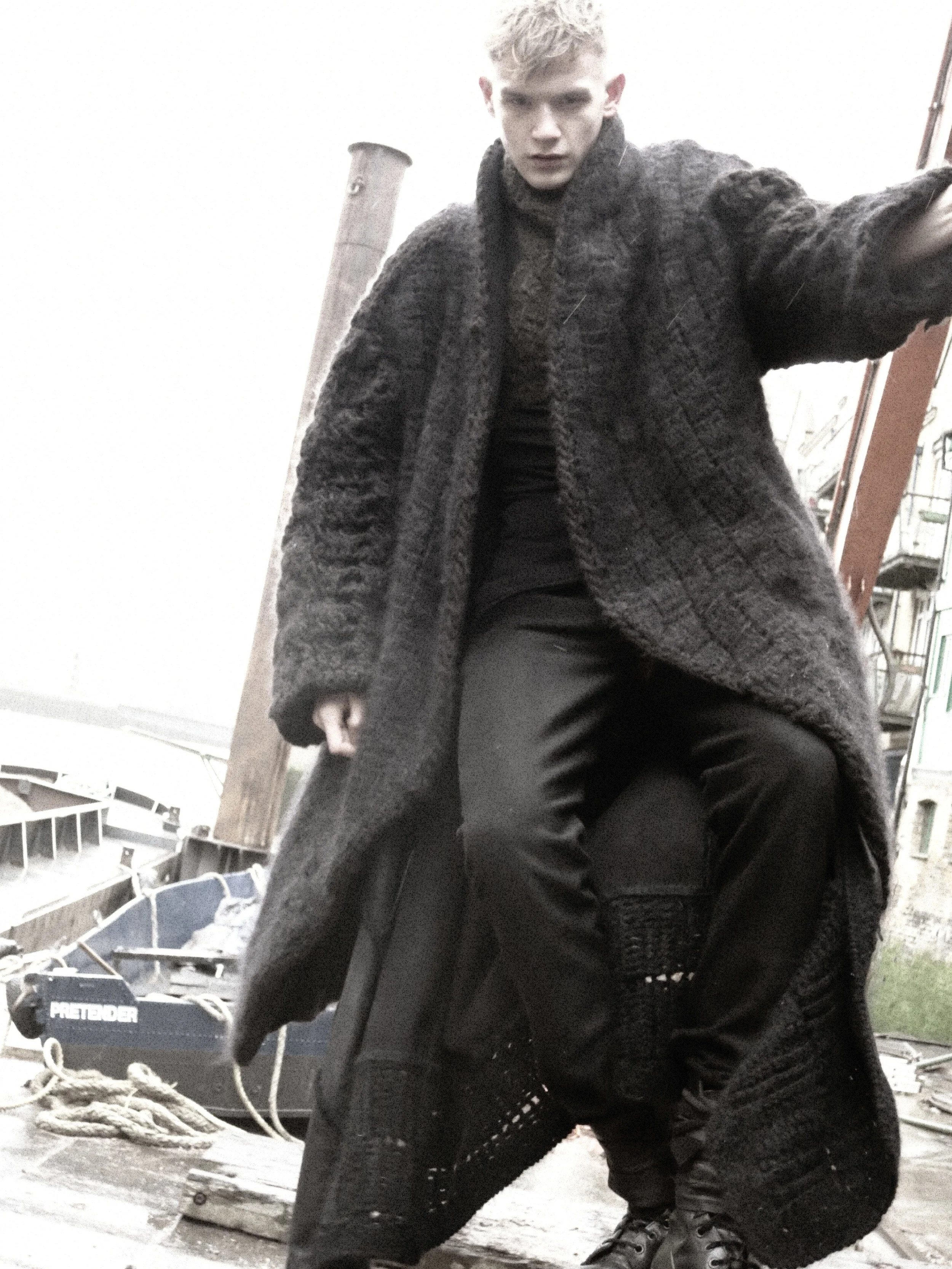 Young man with blond hair wearing a dark knitted sweater, crouching outdoors near working equipment and other materials.