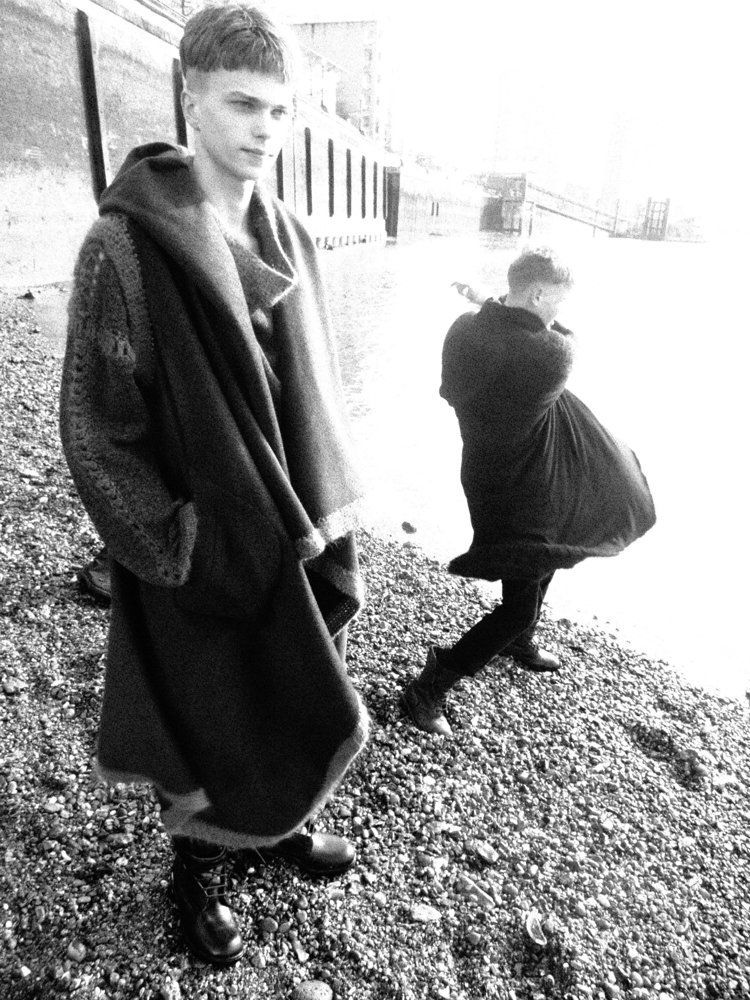 Two boys standing on a gravel surface outdoors, one in the foreground with hands in pockets wearing a long coat, and one in the background with a focused expression, wearing a jacket and boots.