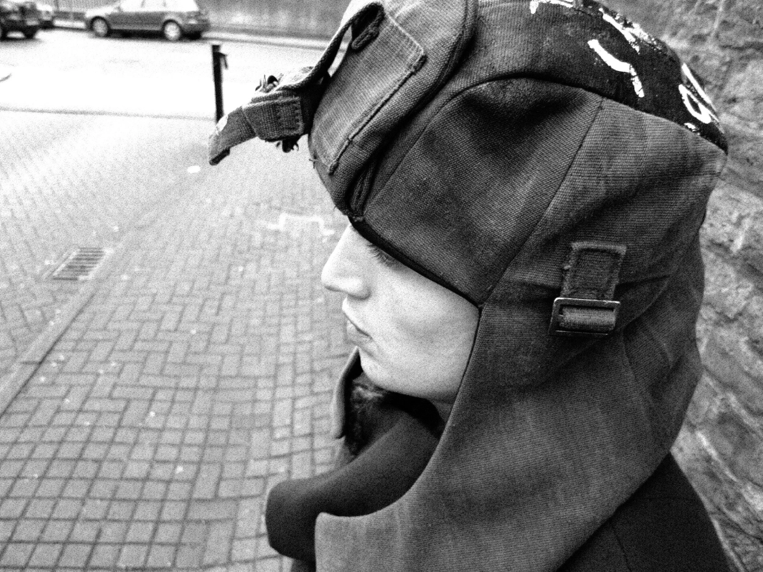 Close-up of a person's head wearing a hat with a strap, standing or walking on a paved sidewalk with parked cars in the background, in black and white.